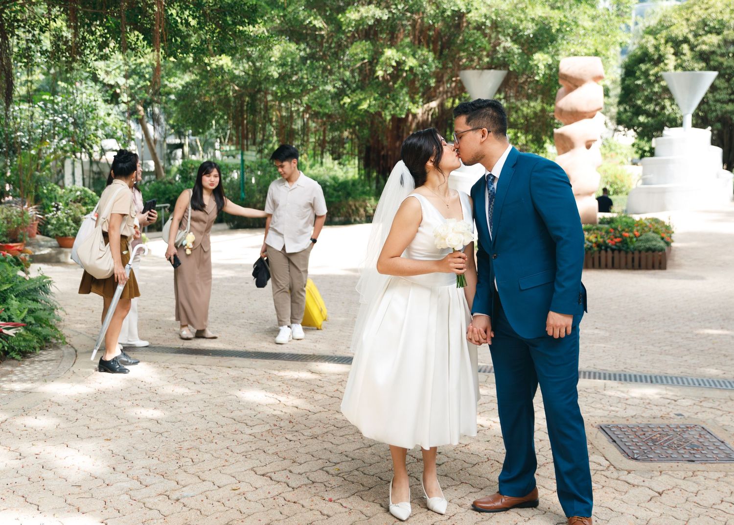 A couple in formal wedding attire walk together through a garden walkway during an outdoor celebration. Civil Ceremony in Hong Kong