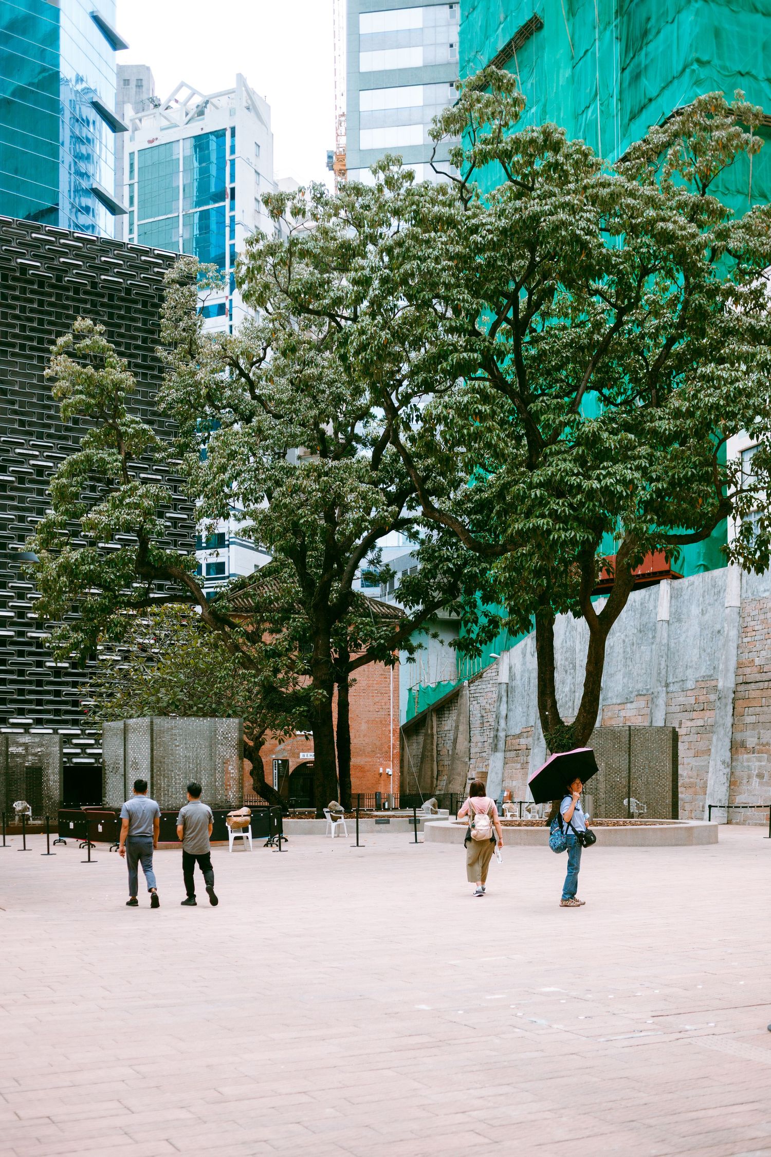 Urban plaza surrounded by modern buildings with green trees and scattered people walking in Hong Kong business district. Taikwun, Hong Kong
