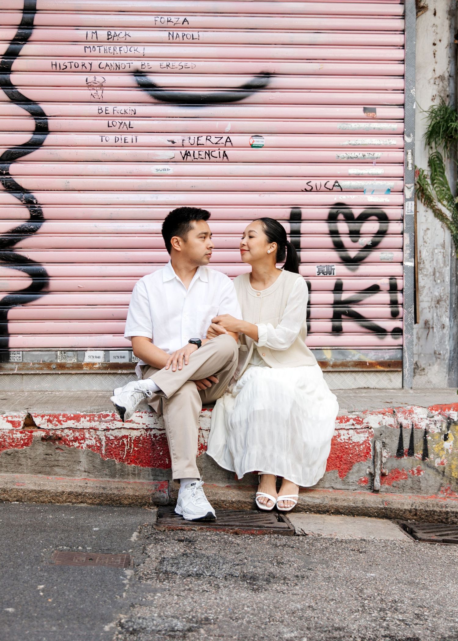 A couple in white outfits sits together on steps against a pink roller shutter door backdrop creating a romantic scene. Couple Photoshoot HK