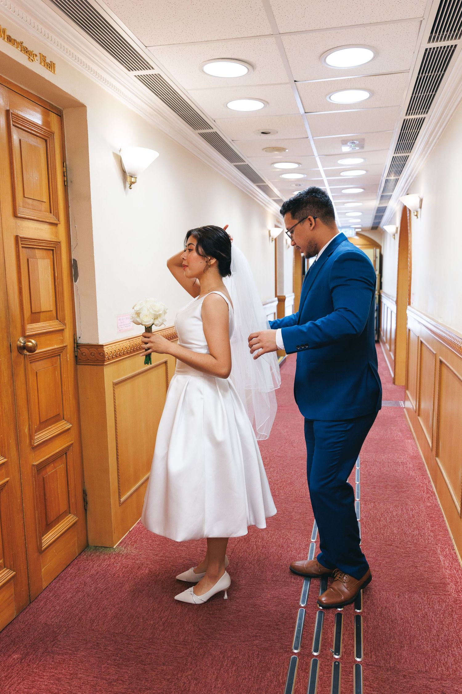 A bride and groom dance playfully in a hallway with red carpet and wooden doors. Civil Ceremony in Hong Kong
