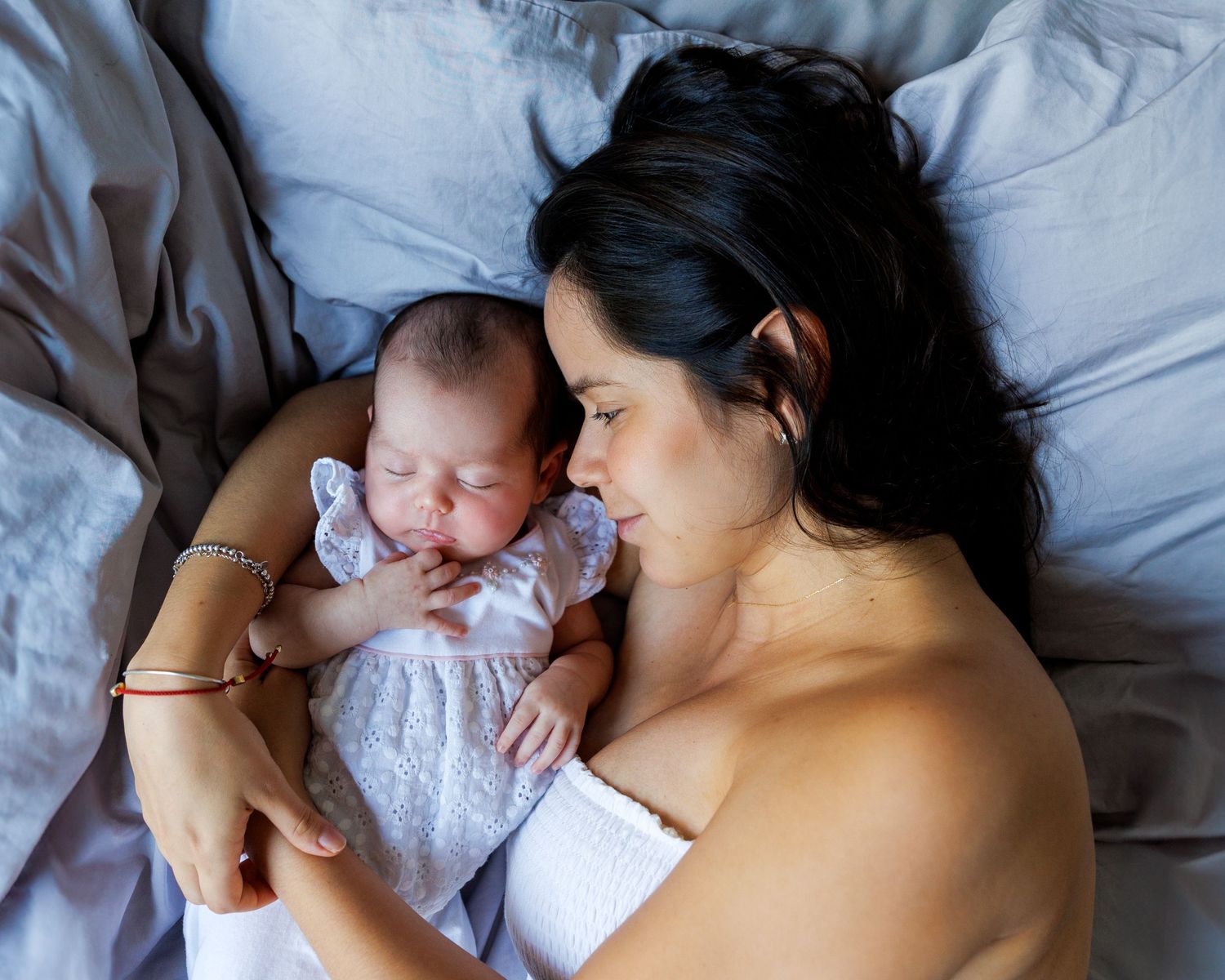 A mother and newborn sharing intimate skin-to-skin contact while lying together on white bedding. Newborn Photographer in Hong Kong