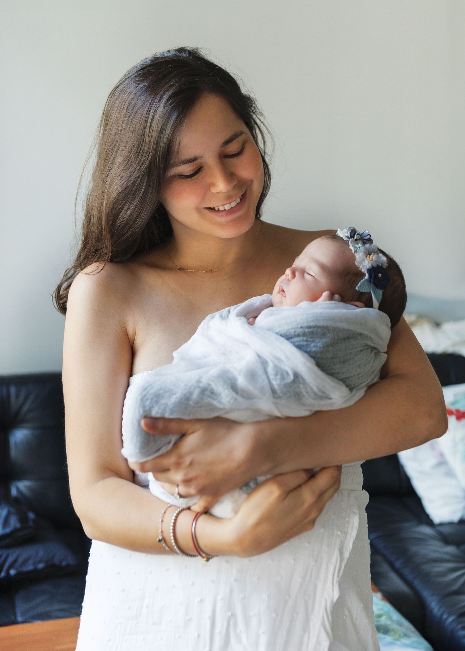 A woman in grey holds a swaddled newborn baby while sitting on a dark couch, shown in a series of tender moments. Newborn Photographer in HK