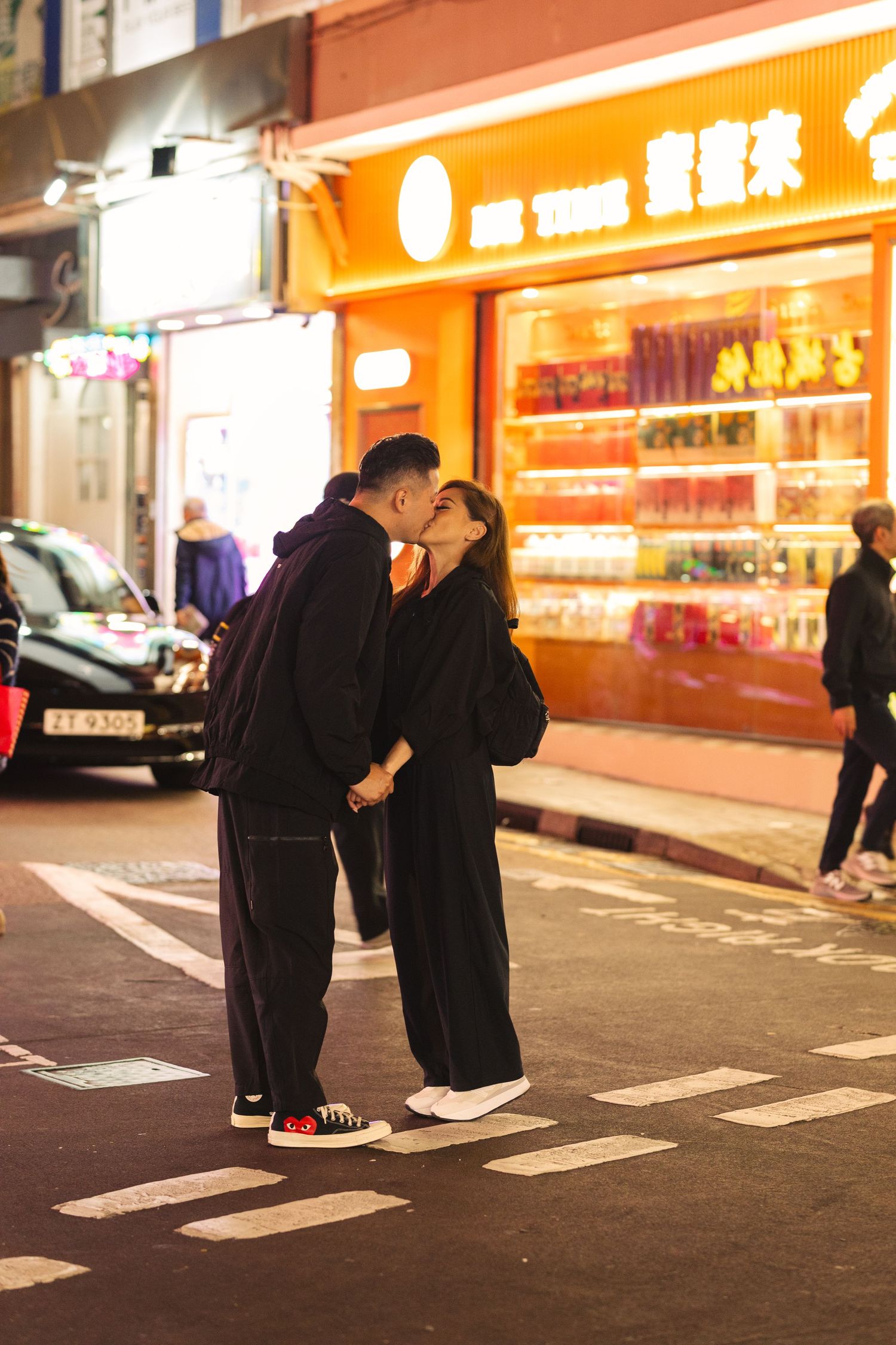 Two figures in dark outfits meet and interact on a city sidewalk at night with glowing storefront signs in the background. Couple Photoshoot