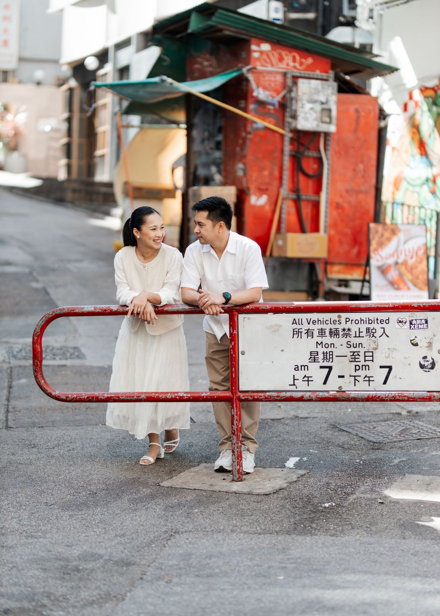A couple dressed in white clothes sits playfully on a metal barrier in an urban street setting. Couple Photoshoot in Central, Hong Kong