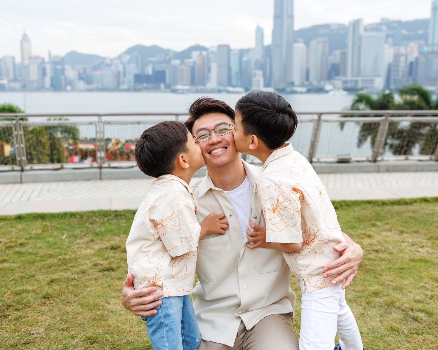 A person wearing glasses receives kisses on cheeks at a scenic overlook of Hong Kong's Victoria Harbour skyline. Hong Kong Family Photograph