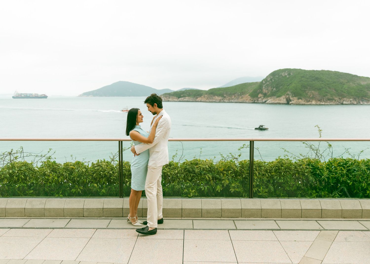 A romantic couple in white clothing shares a tender kiss on a waterfront promenade with coastal hills in the background. Couple Photoshoot