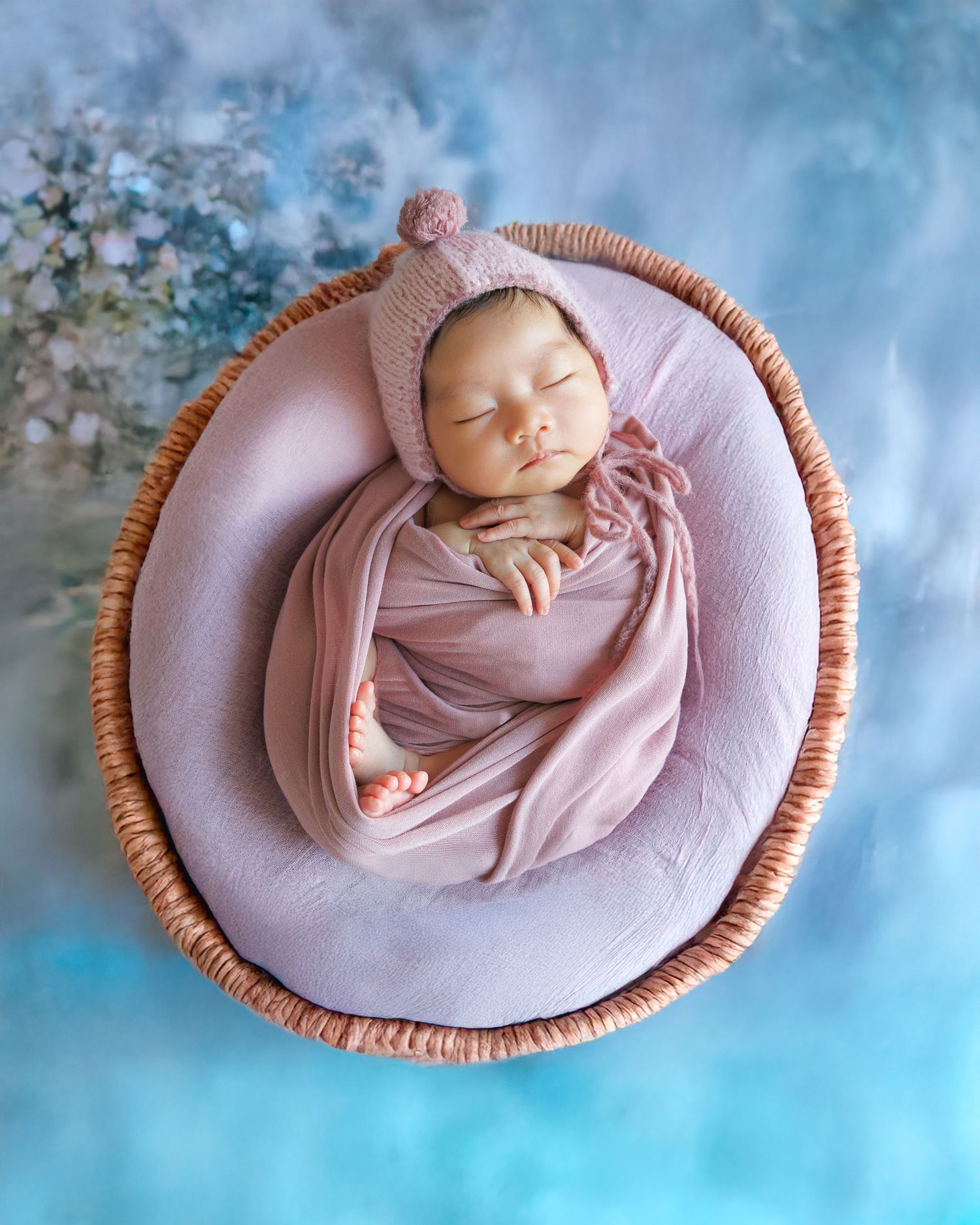 A sleeping newborn baby wrapped in soft pink fabric lies peacefully in a round wicker basket against a blue background. Newborn Photography