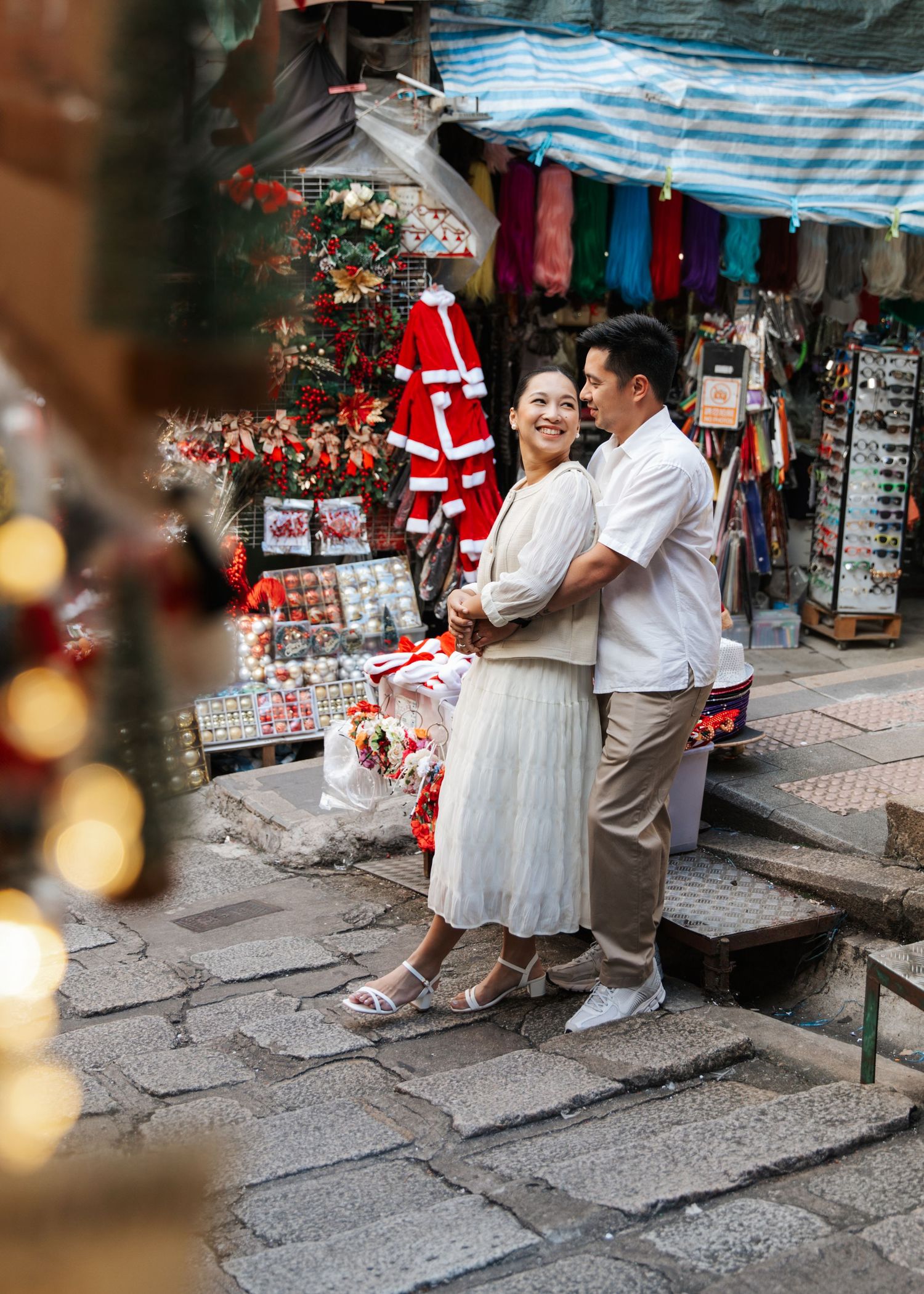 A couple on stone steps near a street market with striped awnings and festive decorations. Couple Photoshoot in Central, Hong Kong
