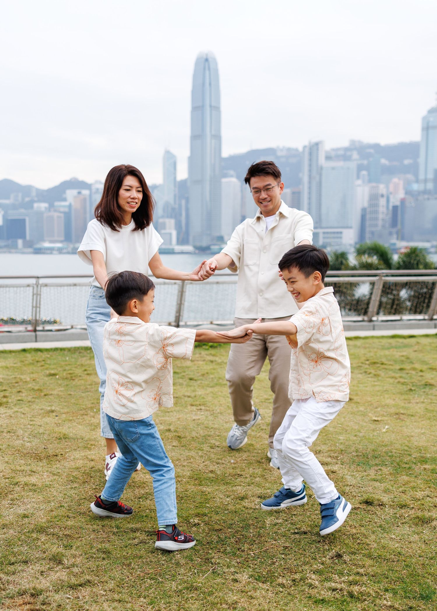 A happy family playing and dancing together on grass with Hong Kong's iconic skyline and harbour view in the background. Hong Kong Photos
