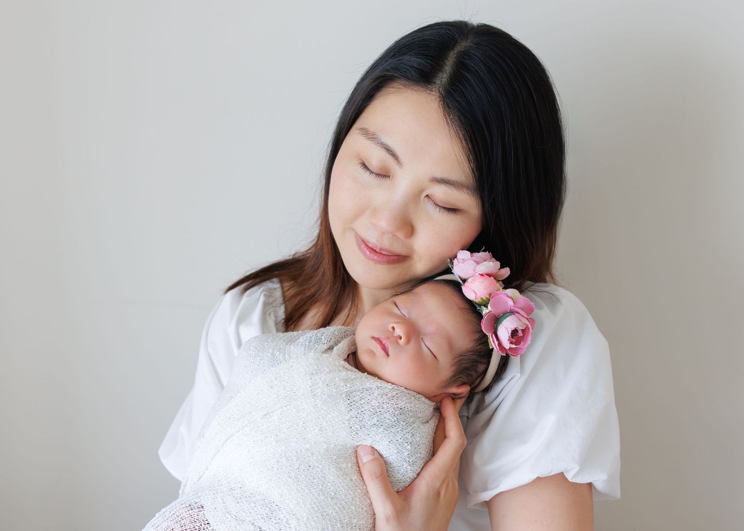 Mother in white dress holds sleeping newborn baby adorned with pink floral headband. Newborn Photography in Hong Kong
