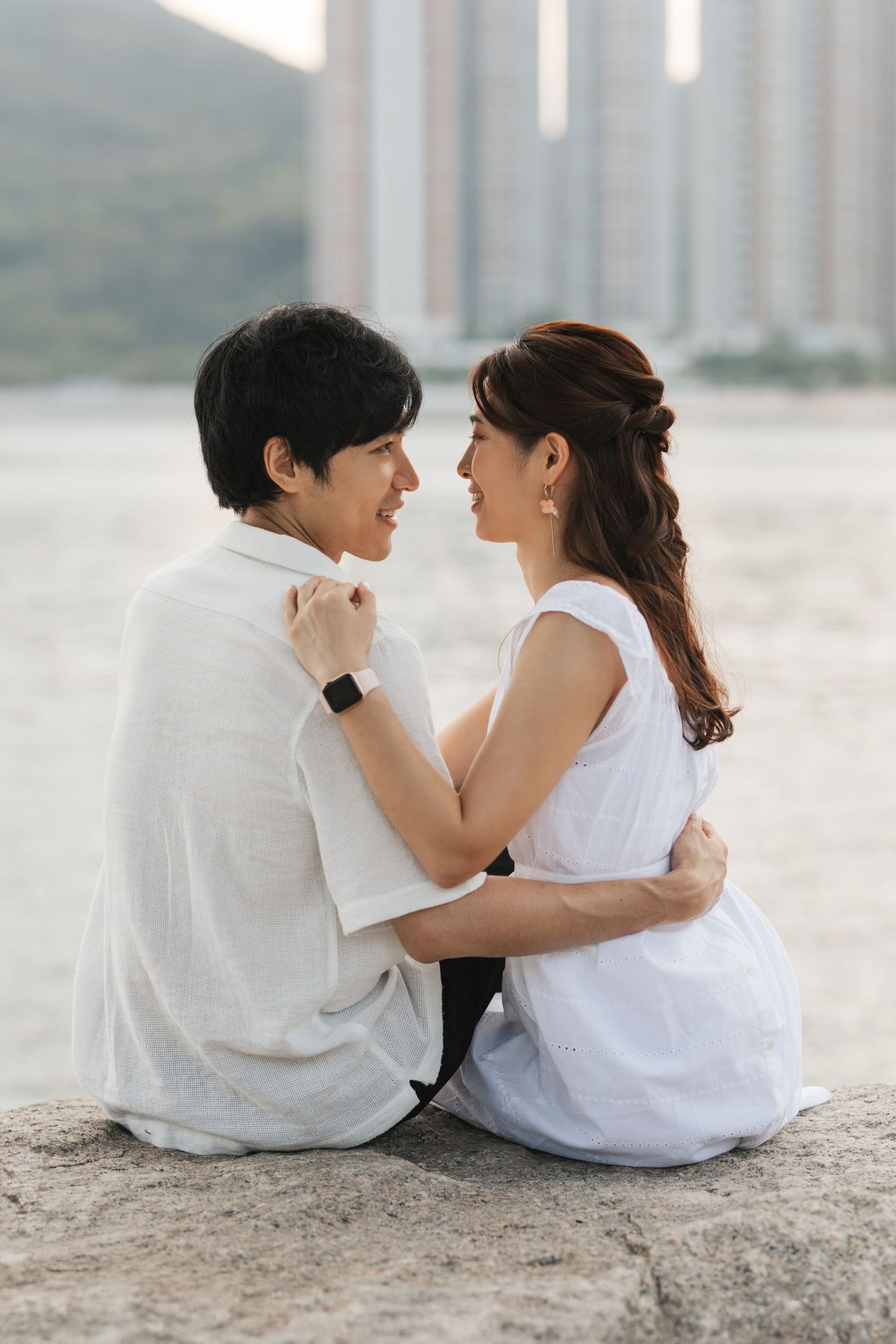 Romantic couple in white clothing sitting close together by the water during a tender moment at sunset. Pre Wedding in Hong Kong