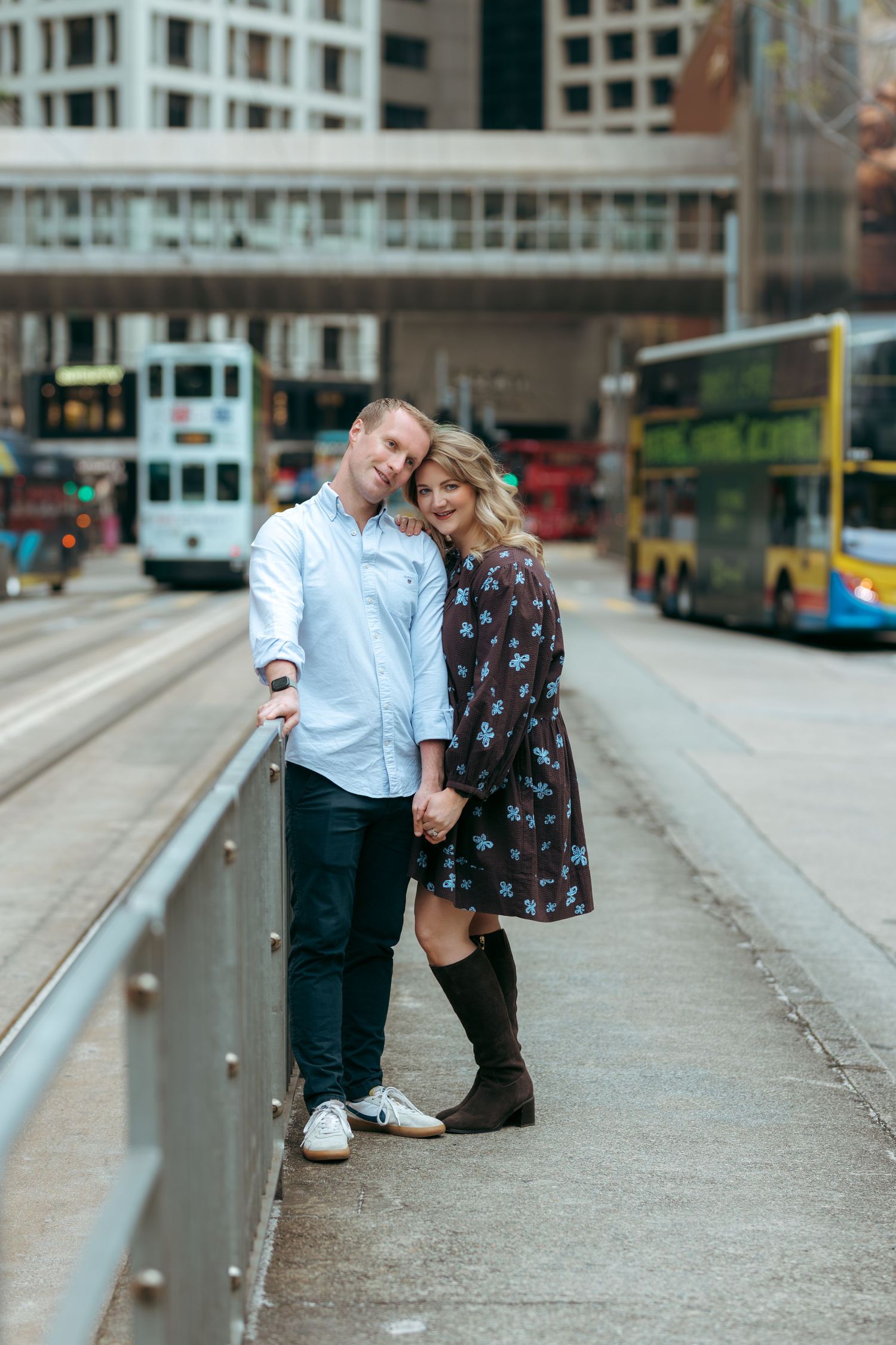 Couple posing on urban street with trams and buses in background, woman in floral dress and boots, man in blue shirt. couple session in HK