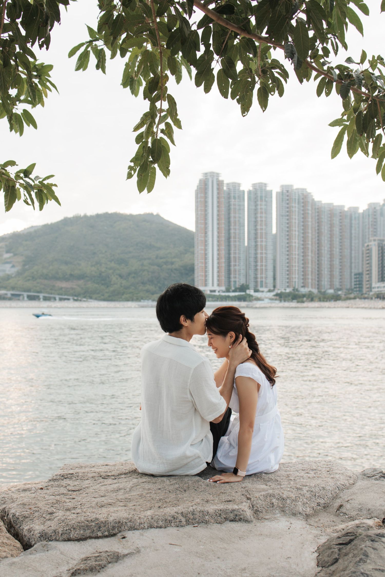 Romantic couple sitting on a rocky shoreline with high-rise buildings and mountains visible across the water. Couple Photoshoot in Hong Kong