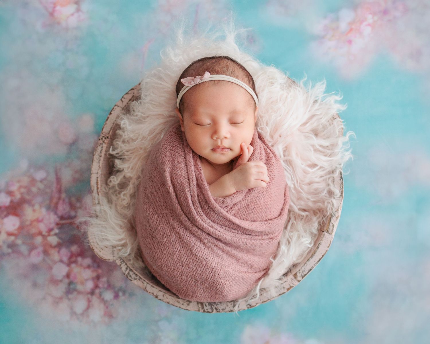 A sleeping newborn wrapped in a pink swaddle rests in a white round bowl against a dreamy blue and pink backdrop. In-home newborn session HK
