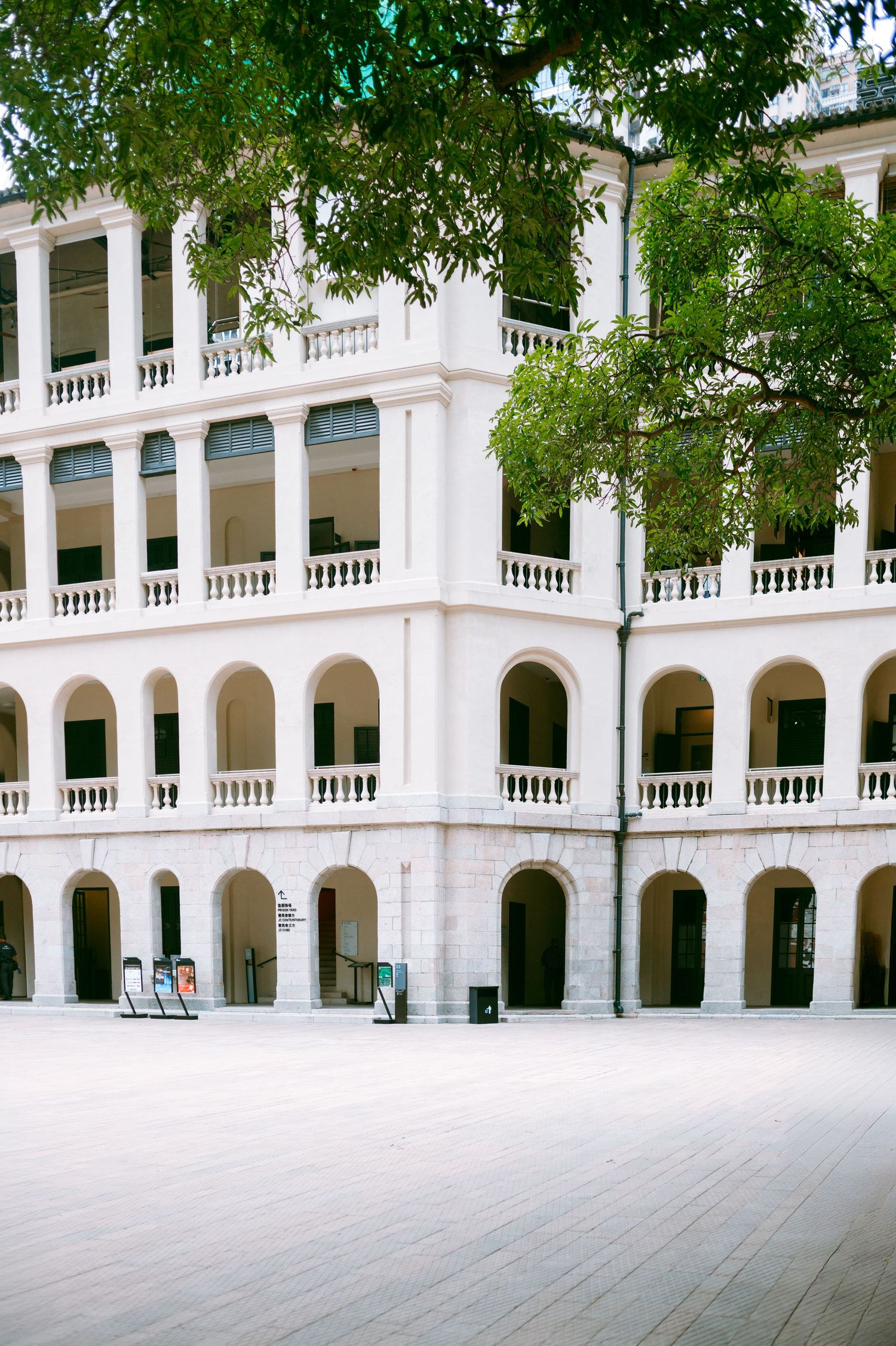A white neoclassical building with a colonnade walkway and arched openings features elegant balustrades and green trees. Taikwun, Hong Kong