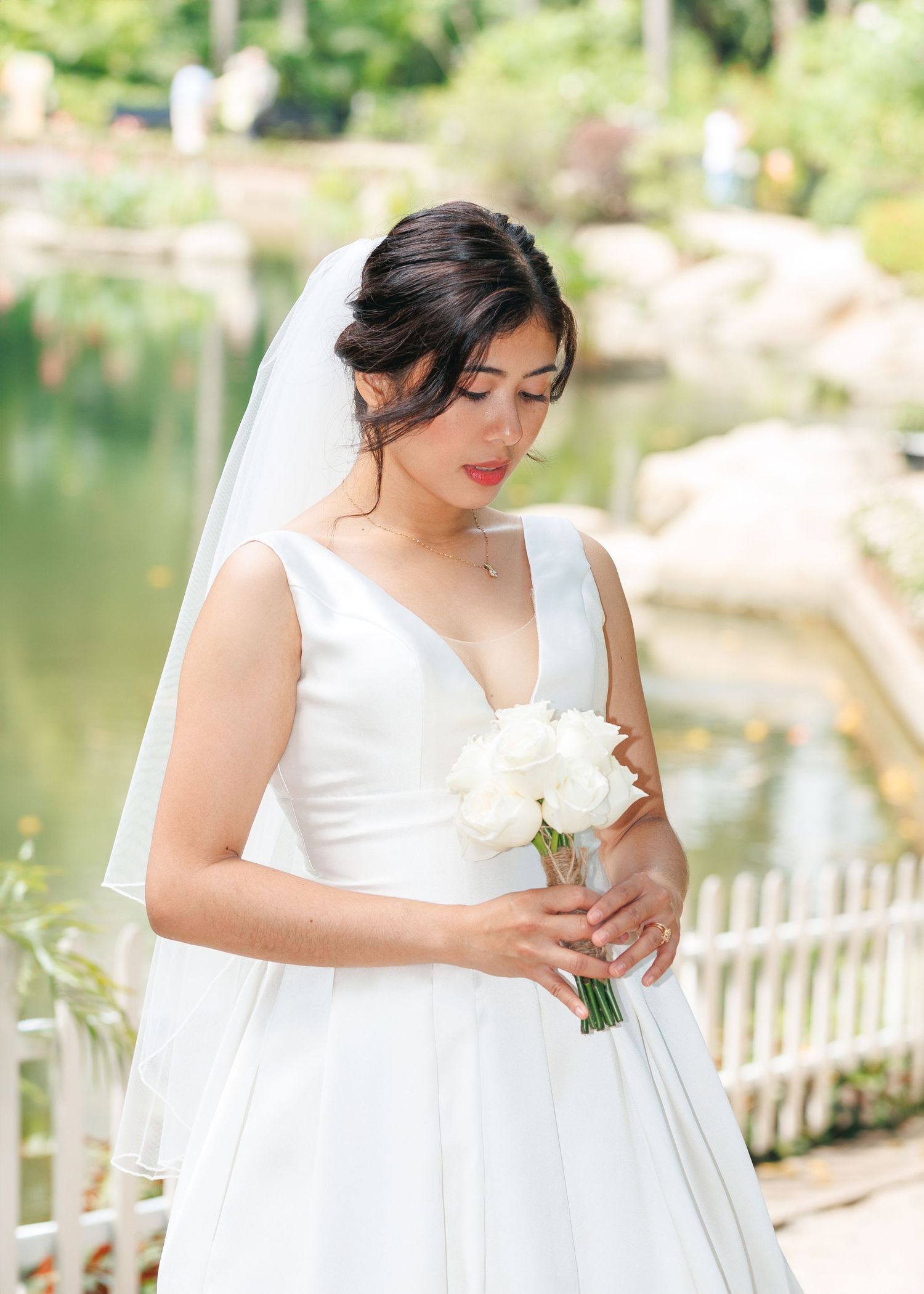 Bride in white wedding dress holding flowers poses gracefully by a lakeside fence in a garden setting. Civil Ceremony in Hong Kong