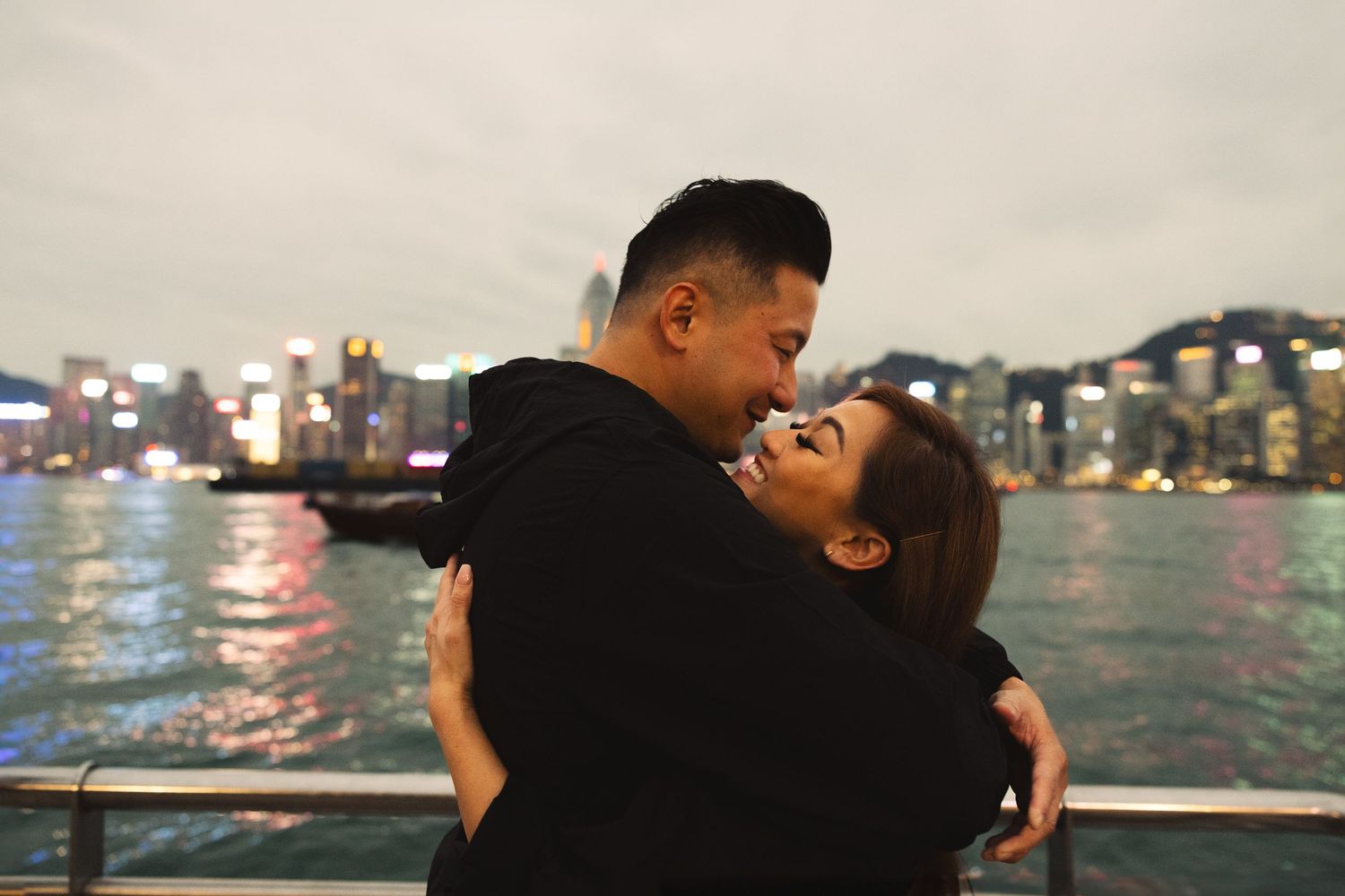 A loving couple embraces and laughs together against the illuminated Hong Kong waterfront skyline at dusk. Couple Photoshoot in Hong Kong