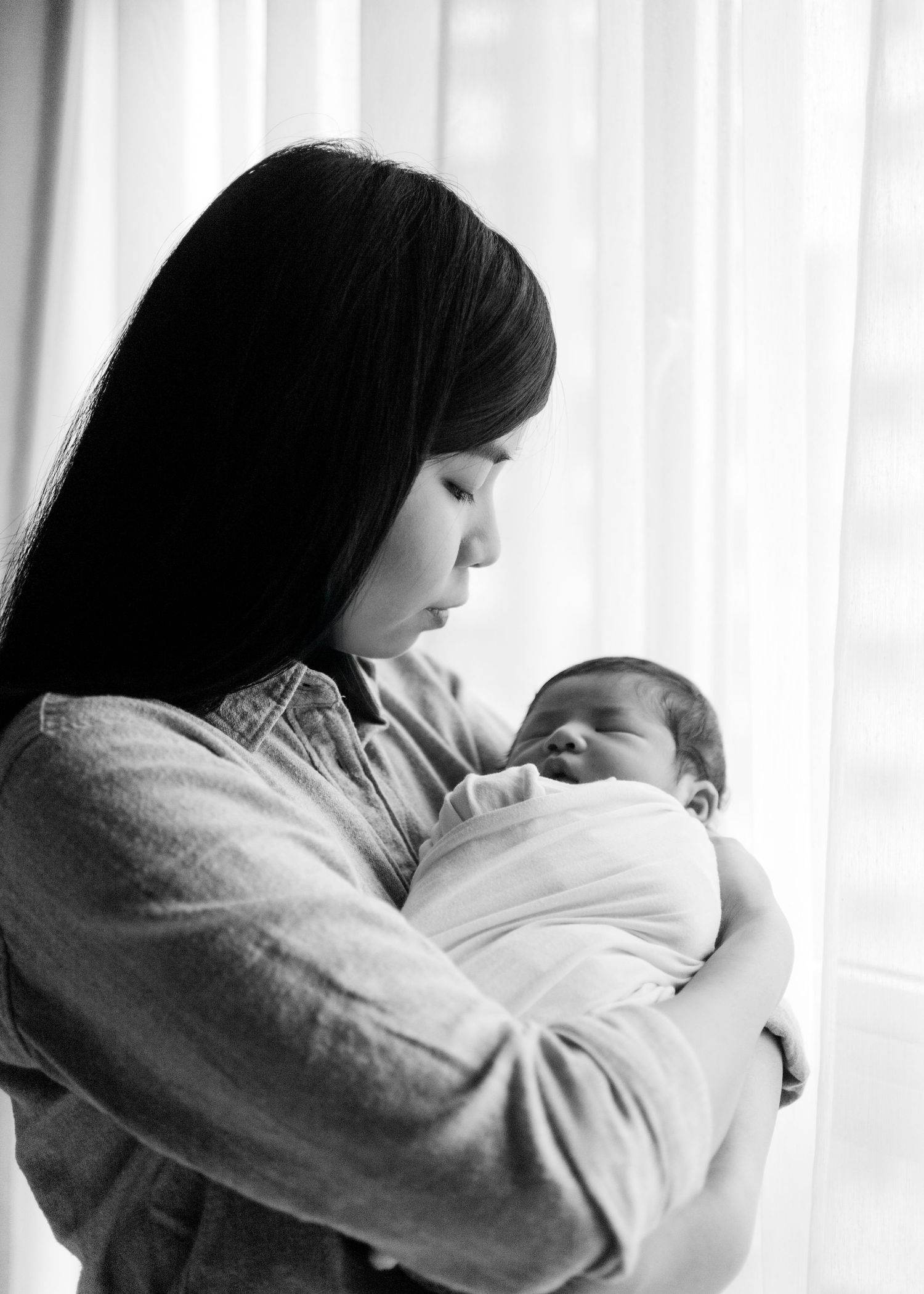 Mother embraces and feeds newborn baby while sitting near window with soft backlighting creating intimate moment. Newborn Photography Hong