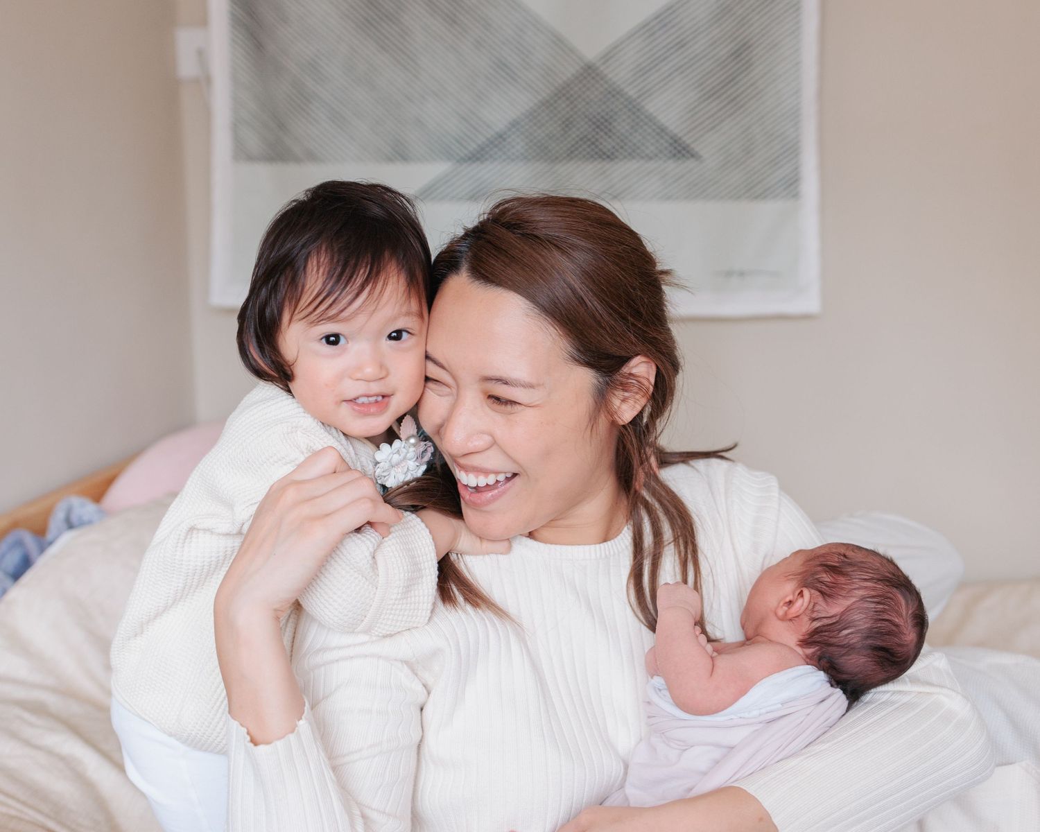 A loving moment on a bed with a mother embracing a toddler while holding a newborn baby, all wearing white. In-Home Newborn Session in HK