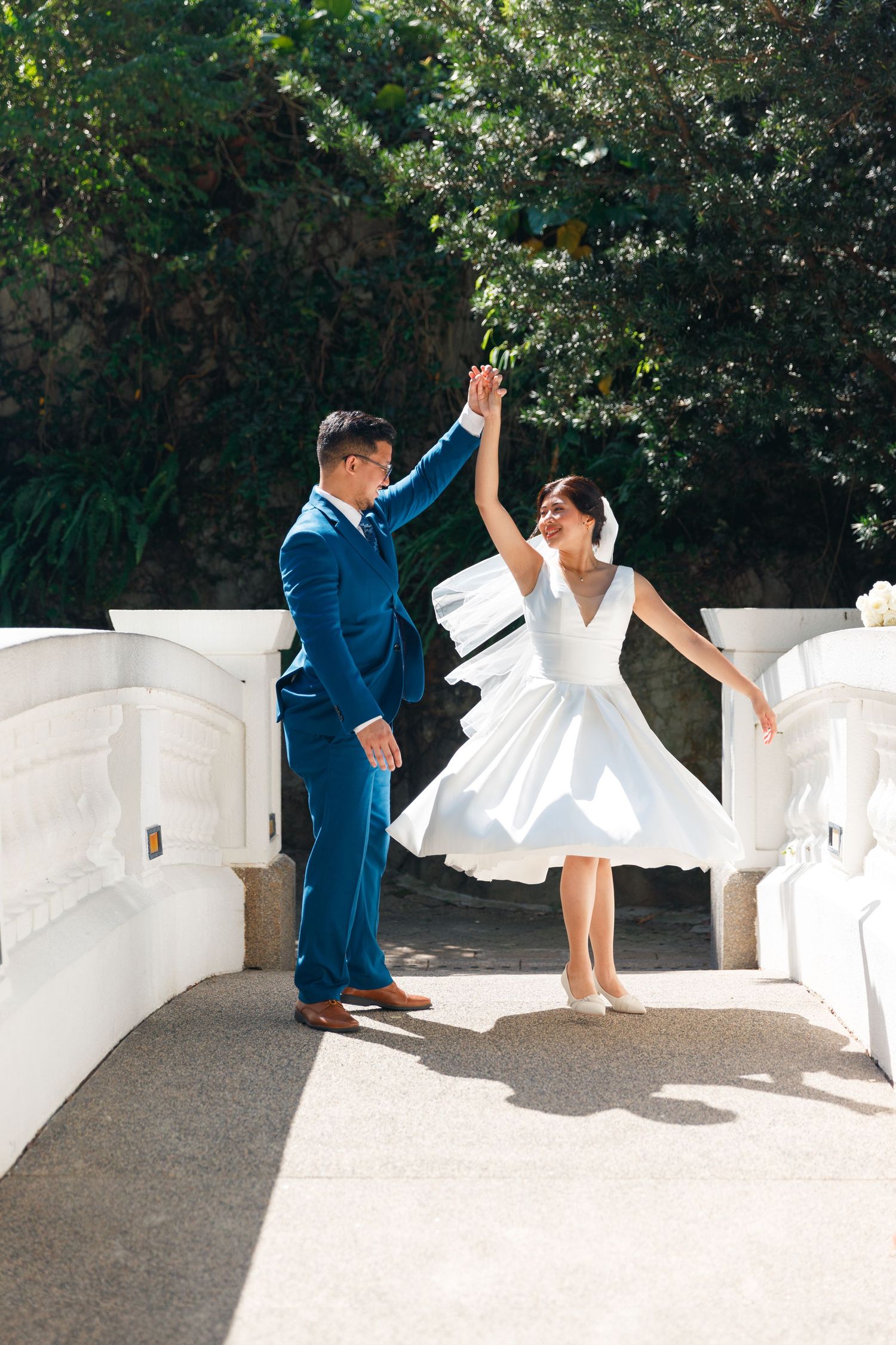 A couple in wedding attire dance and twirl playfully against a white wall backdrop and green foliage. Civil Ceremony in Hong Kong