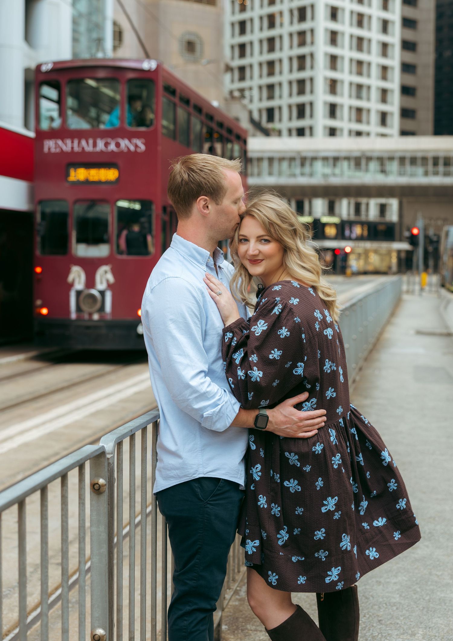 Couple embracing romantically on a city street with a red double-decker Penhaligon's tram passing behind them. couple session in Hong Kong.