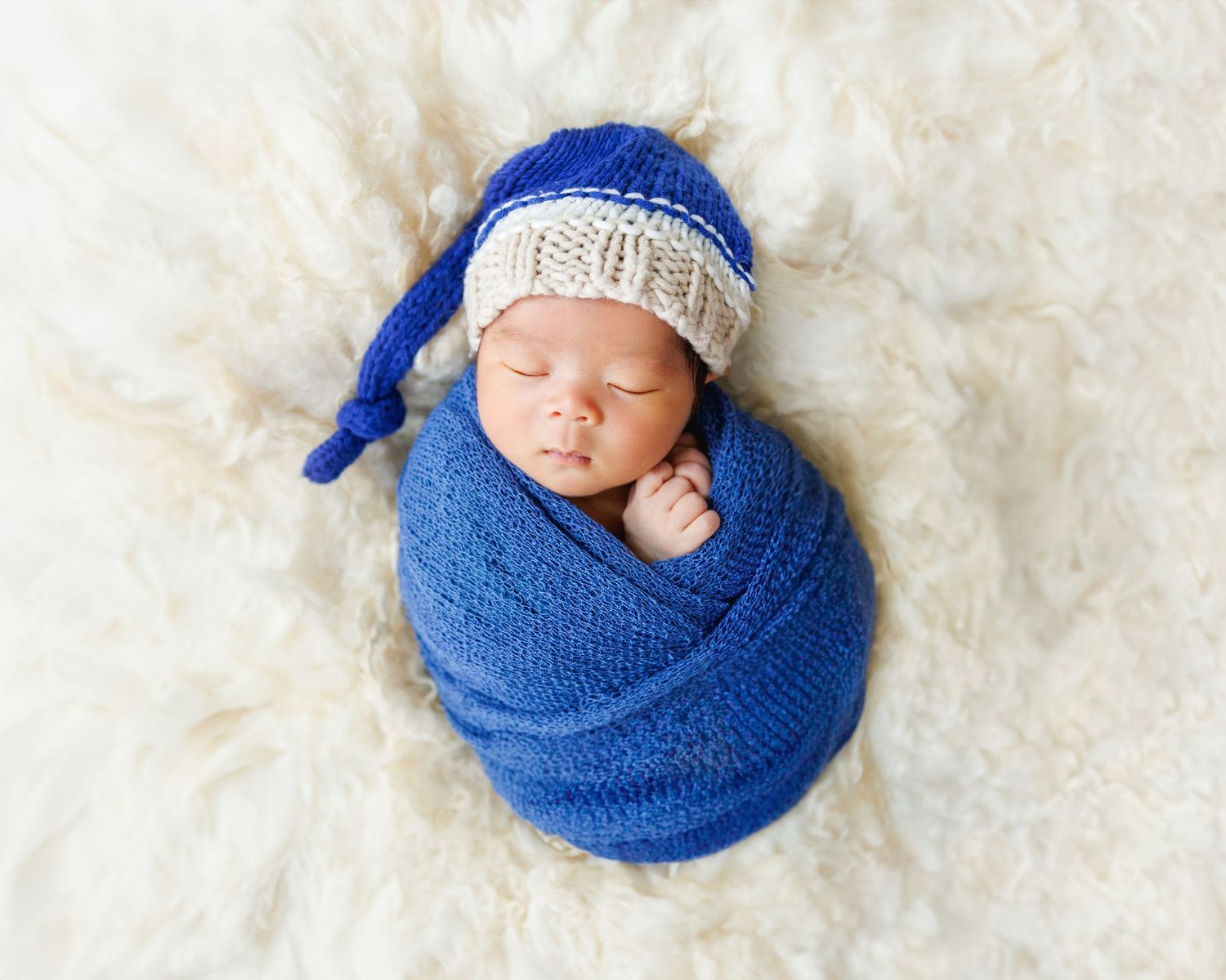A newborn in a blue wrap and knit cap rests peacefully on a plush white surface. Newborn Photographer in Hong Kong