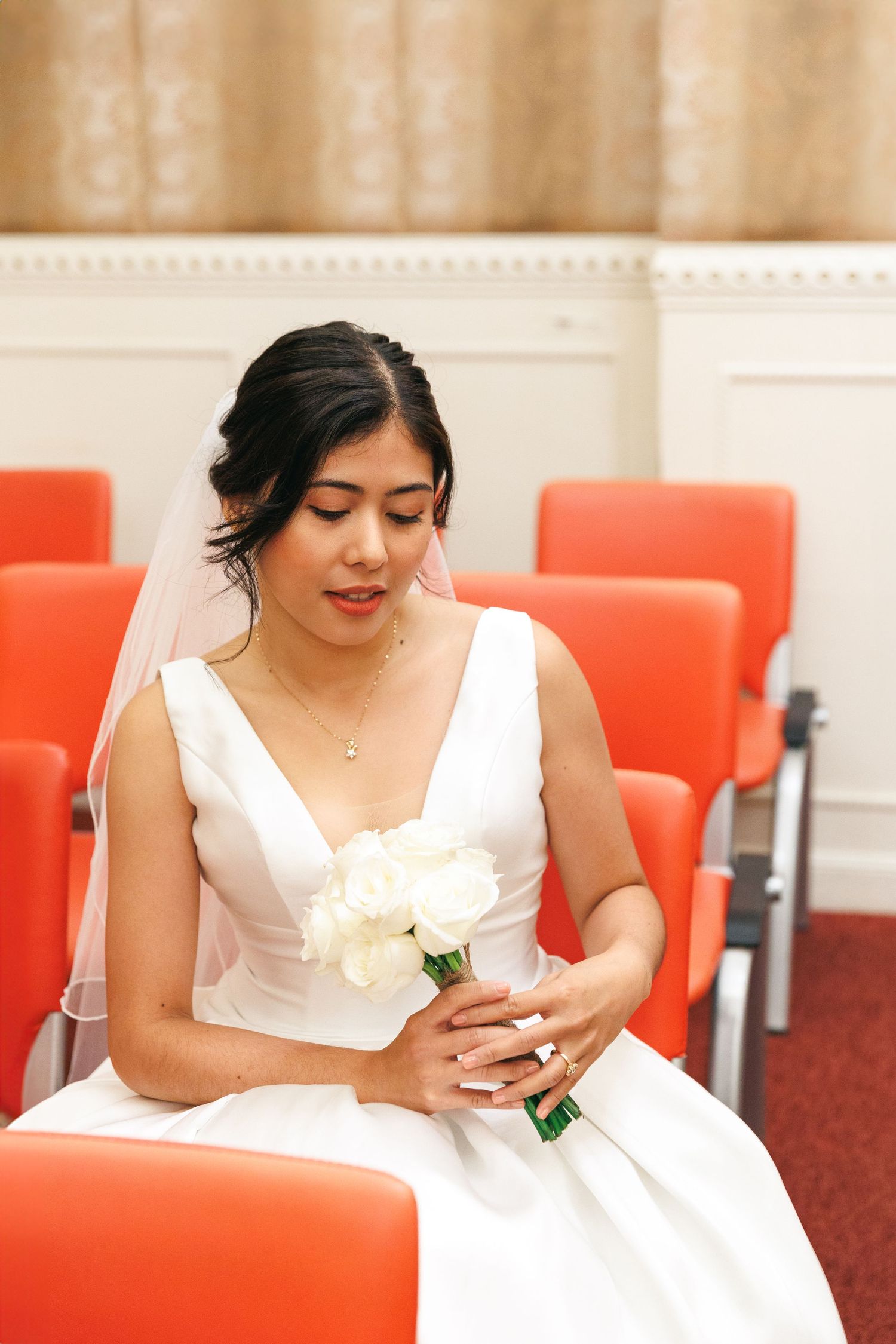 A woman in a white wedding dress sits on an orange chair while looking down at a mobile device. Civil Ceremony in Hong Kong