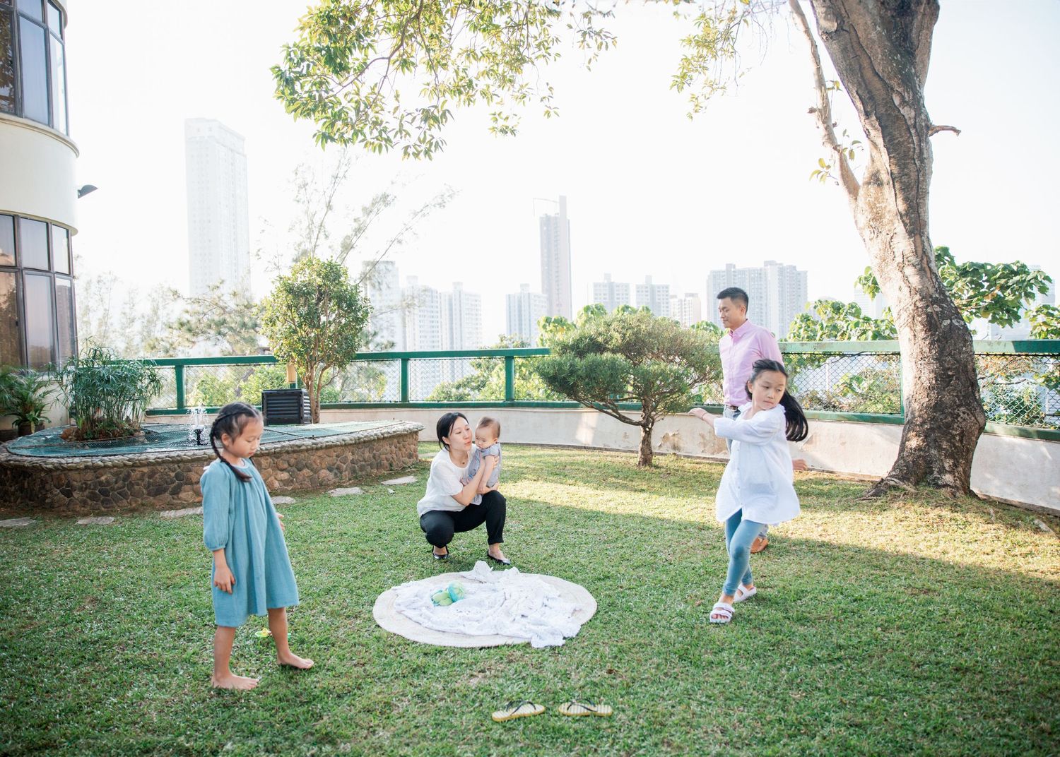 A group plays together on a grassy lawn with a picnic blanket and city buildings visible in the background. Hong Kong Family Photographer
