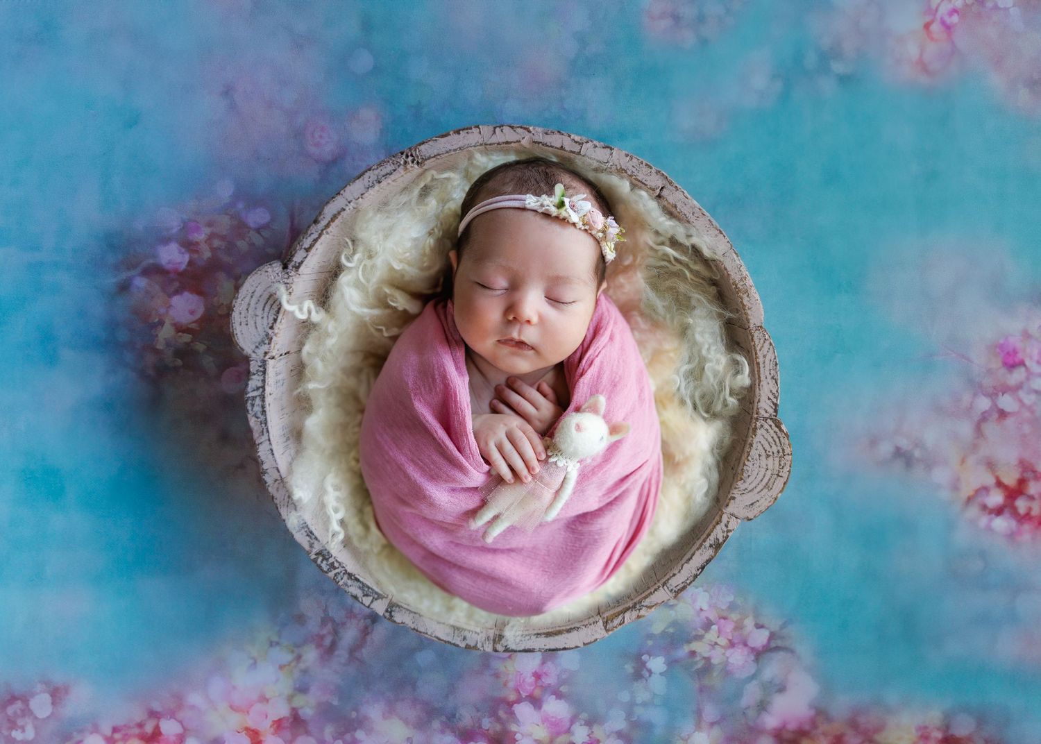 A baby wrapped in pink fabric lies peacefully on a textured bowl prop against a turquoise and pink floral background. Newborn Photographer