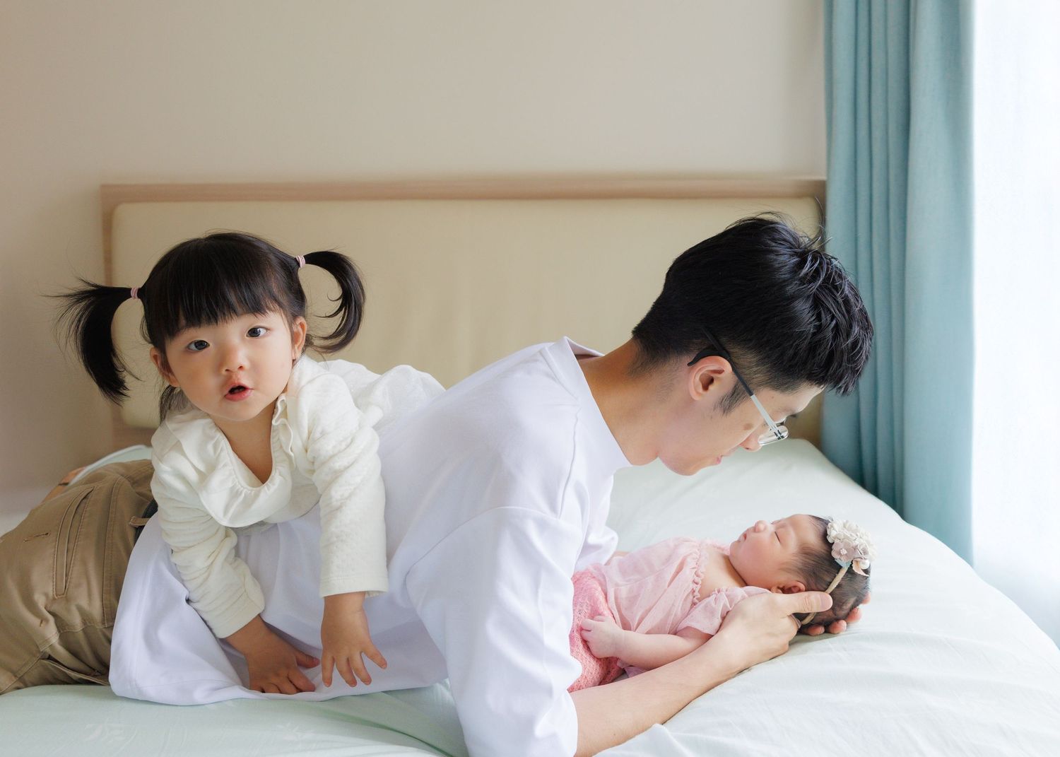 Family relaxes together on white bed with siblings interacting while wearing casual clothes. Newborn Photography in Hong Kong