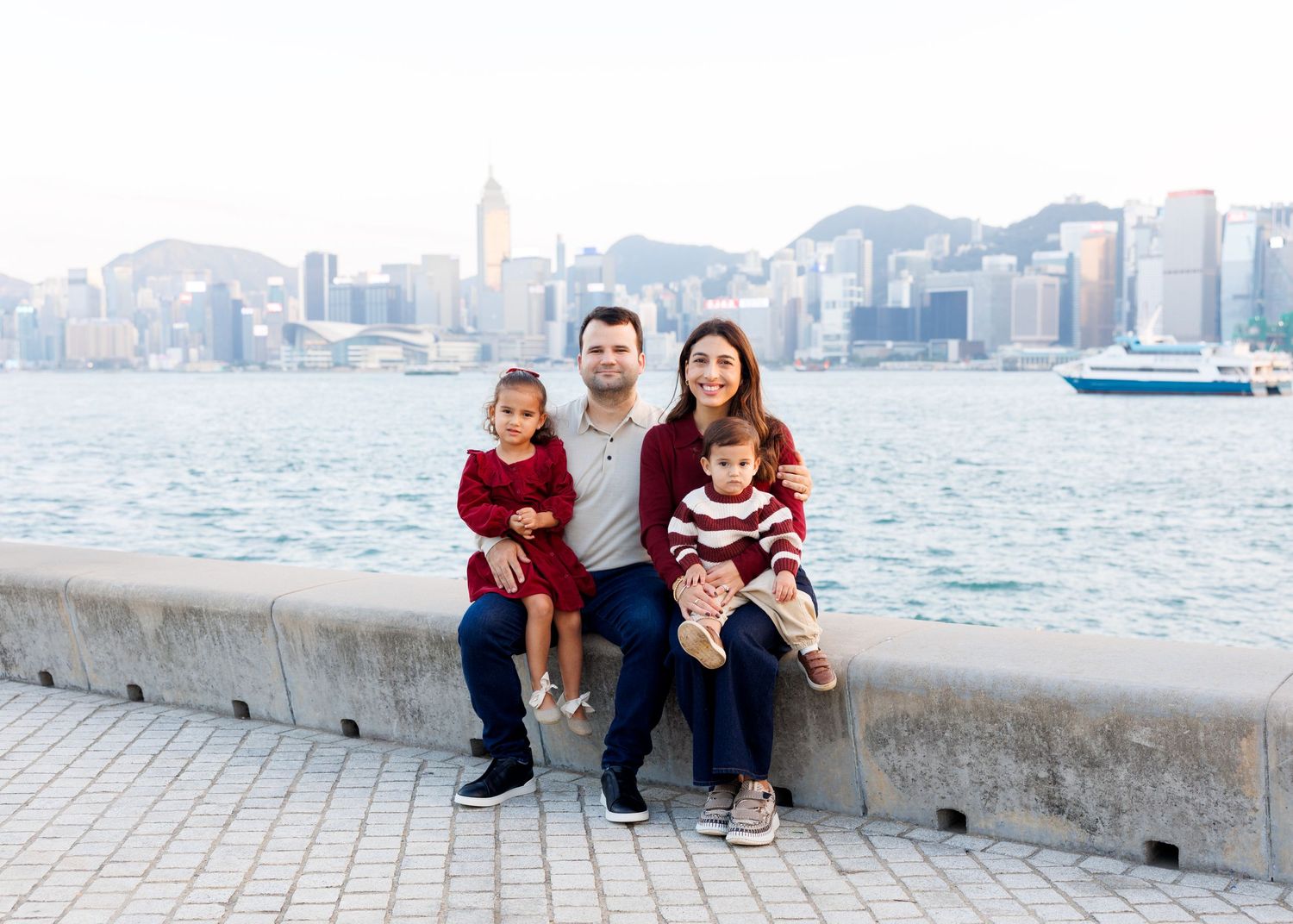 family on a stone ledge overlooking Victoria Harbor with Hong Kong's skyline in the background. Family Photoshoot in Hong Kong