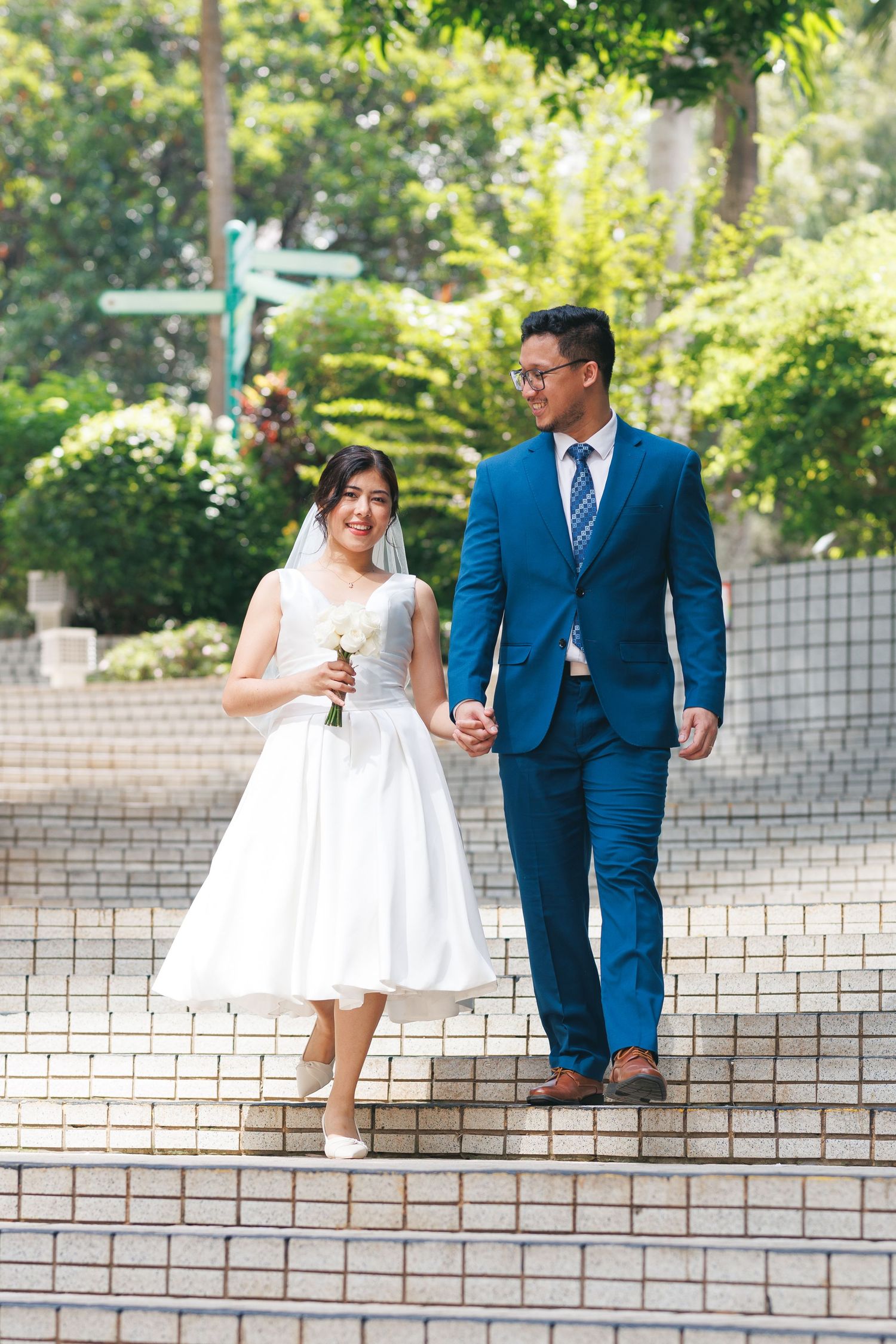 A couple walking up white stone steps outdoors, with a bride in a tea-length dress and groom in a blue suit. Civil Ceremony in Hong Kong