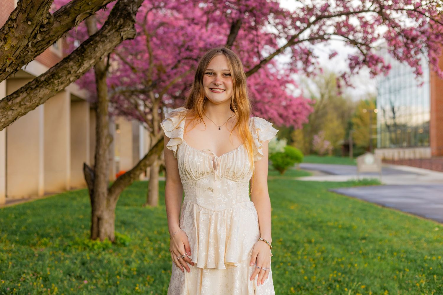 a young woman wearing a white dress stands in front of a pink cherry tree looking at the camera