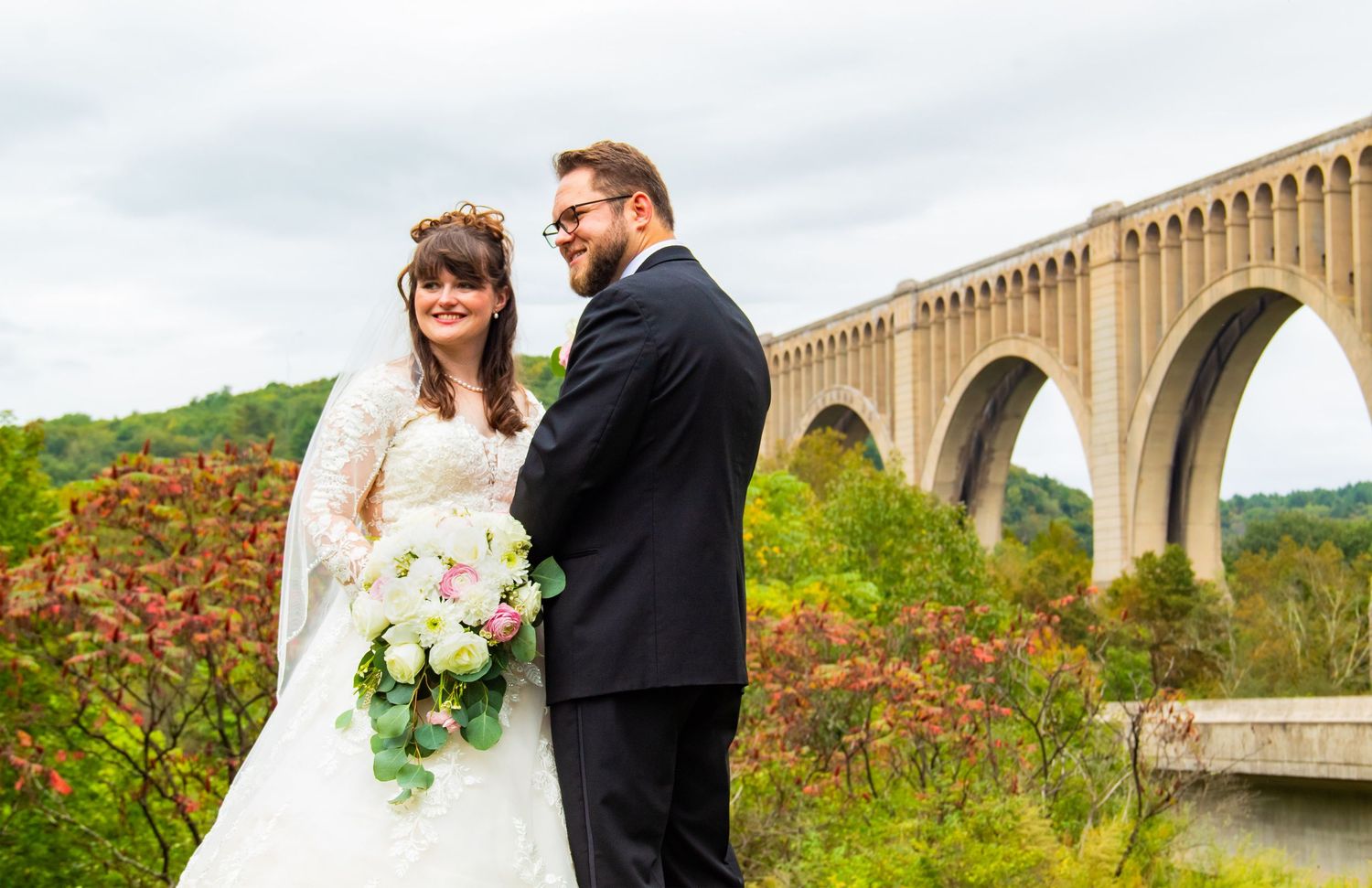 a woman in a wedding dress and a man in a tux stand in front of a bridge looking fondly to the left