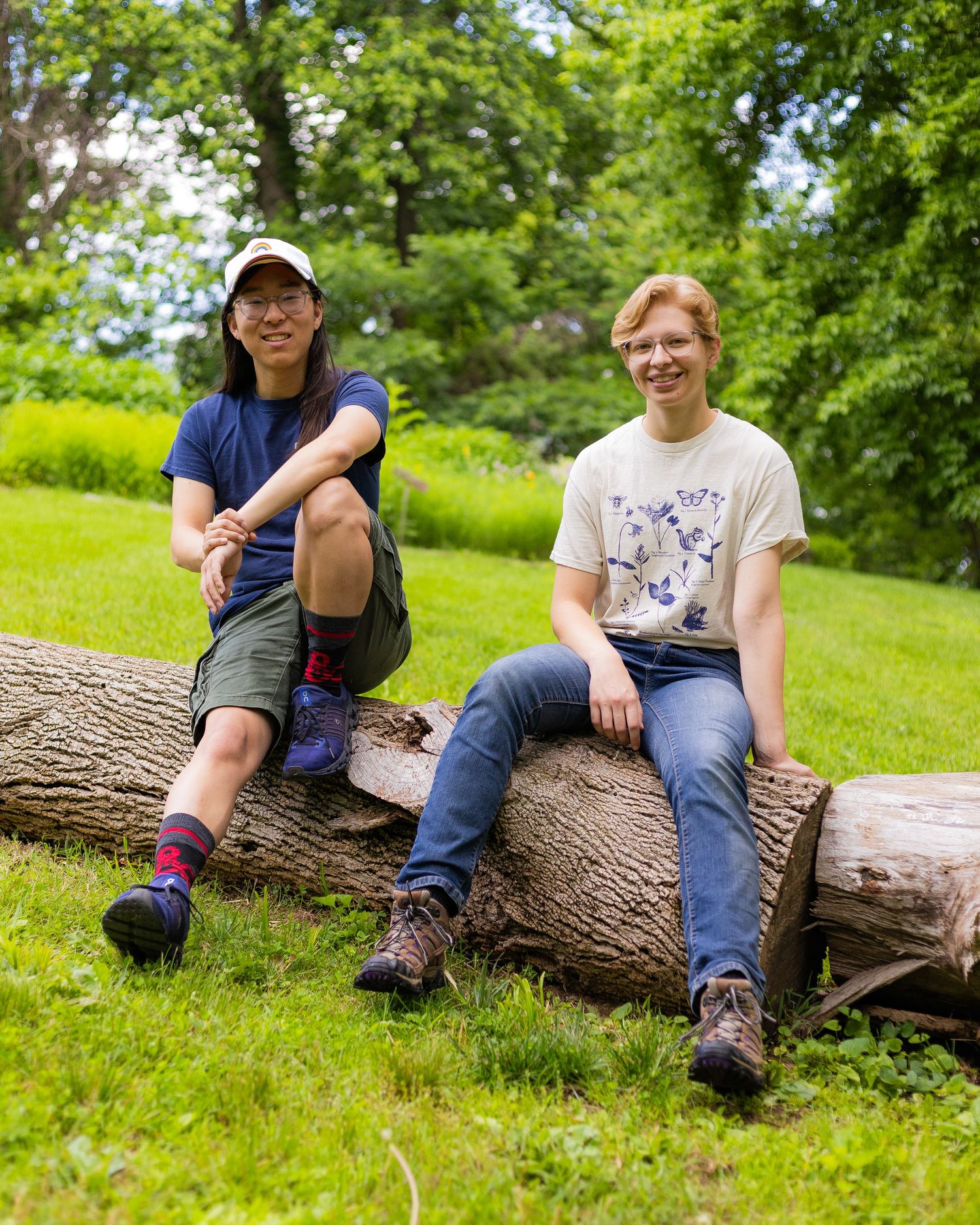 two people sit on a log, posed casually, looking at the camera and surrounded by vibrant green grass and trees