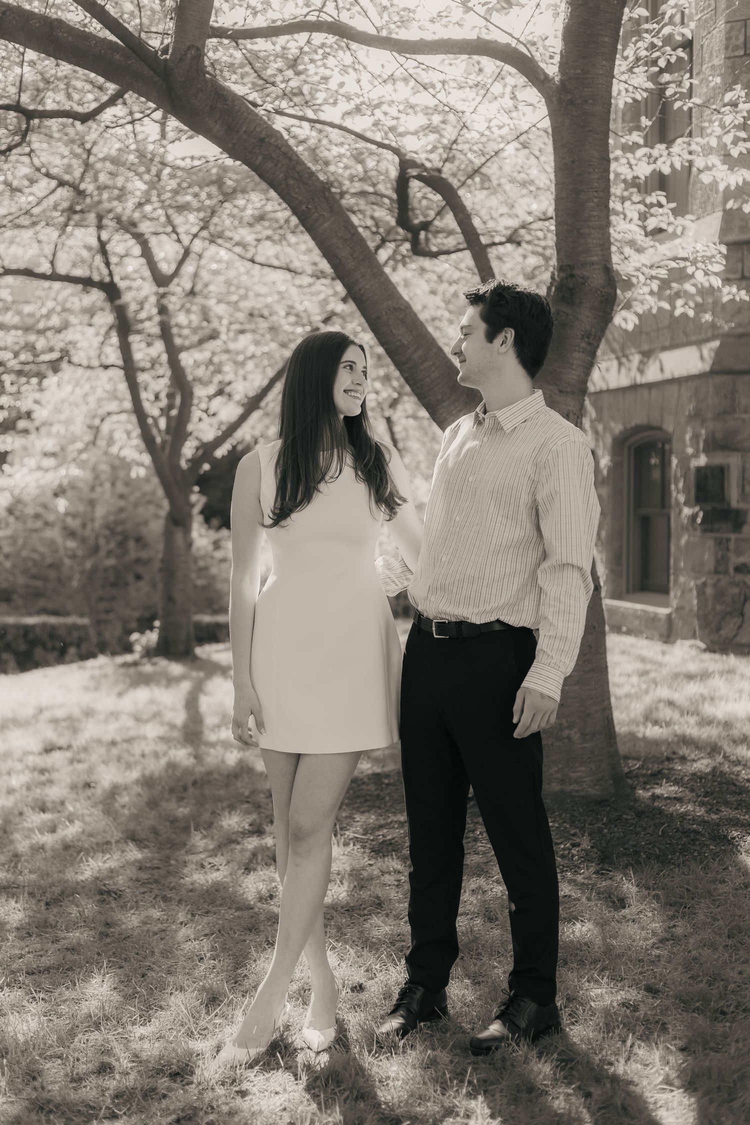 a black and white image. a young couple stand in front of a tree looking at each other and smiling