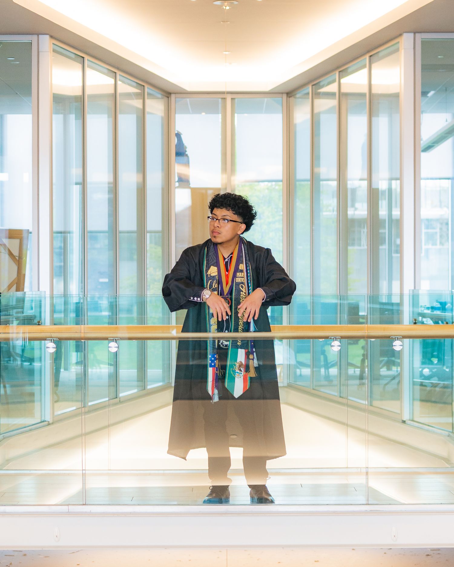 a young man stands leaning up against a railing facing the camera with a brightly lit glass building interior framing him