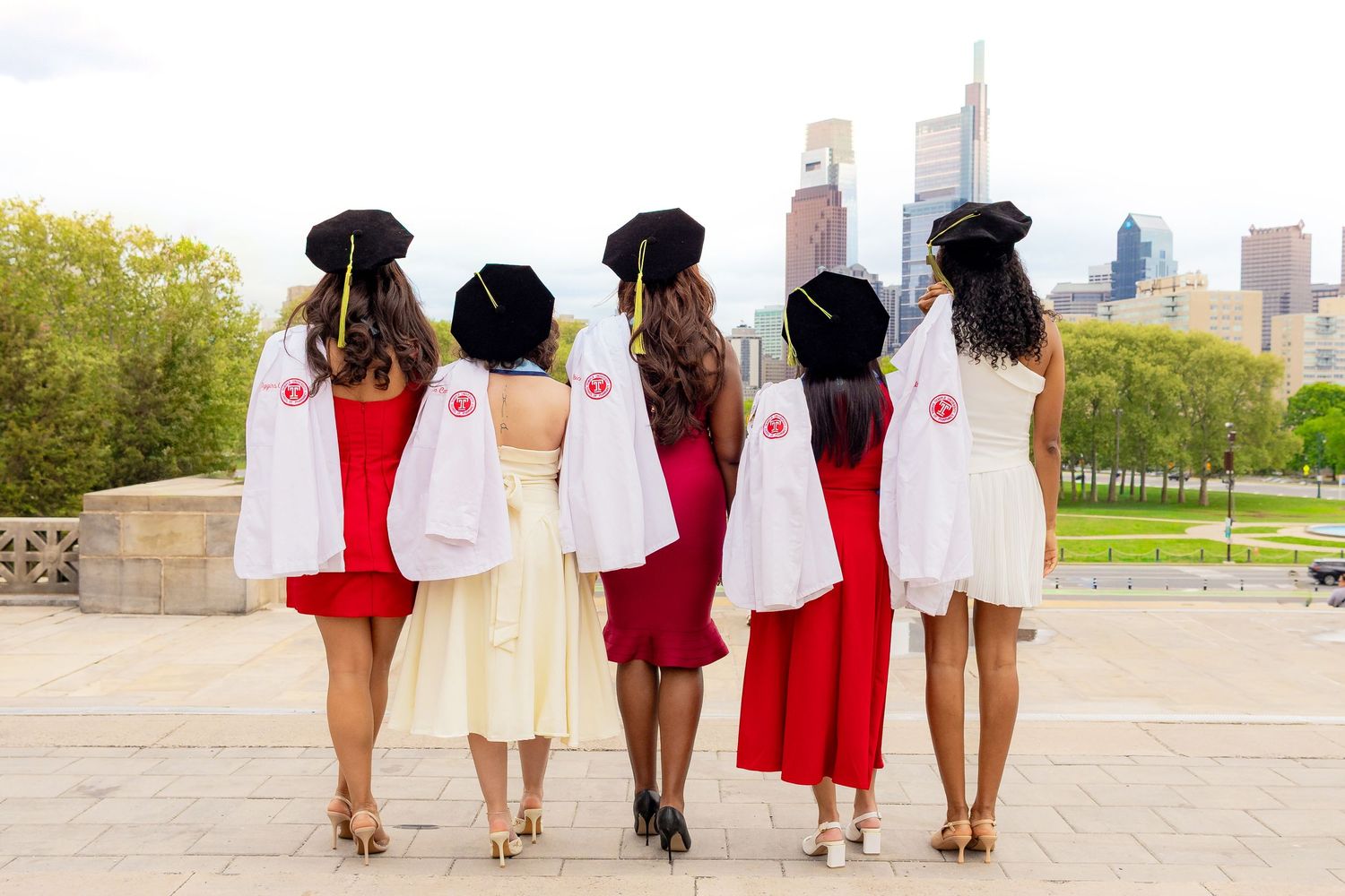 Five women face away from the camera holding pharmacist coats over their left shoulders, looking out over the Philadelphia skyline