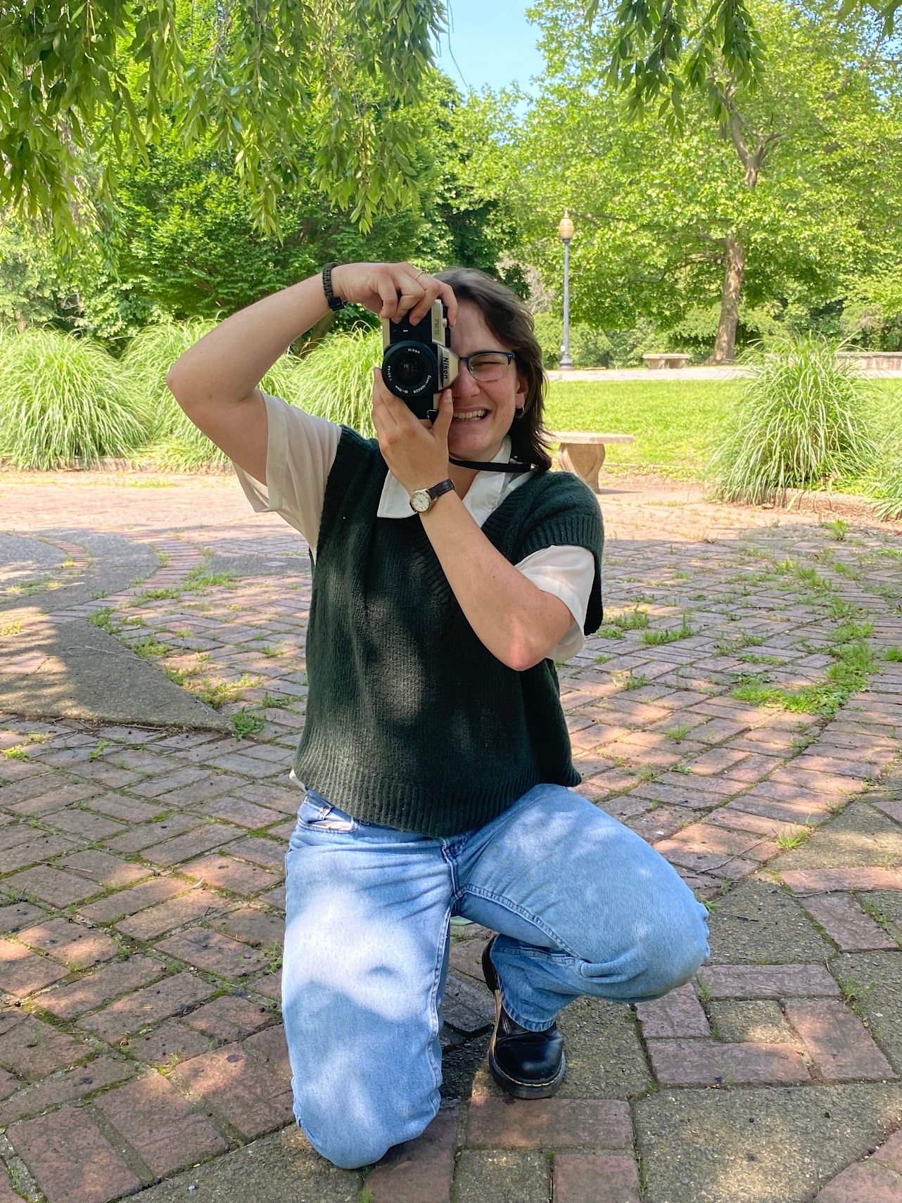 photo of Julie kneeling on the ground in blue jeans and a green sweater vest holding a camera obscuring half of her face