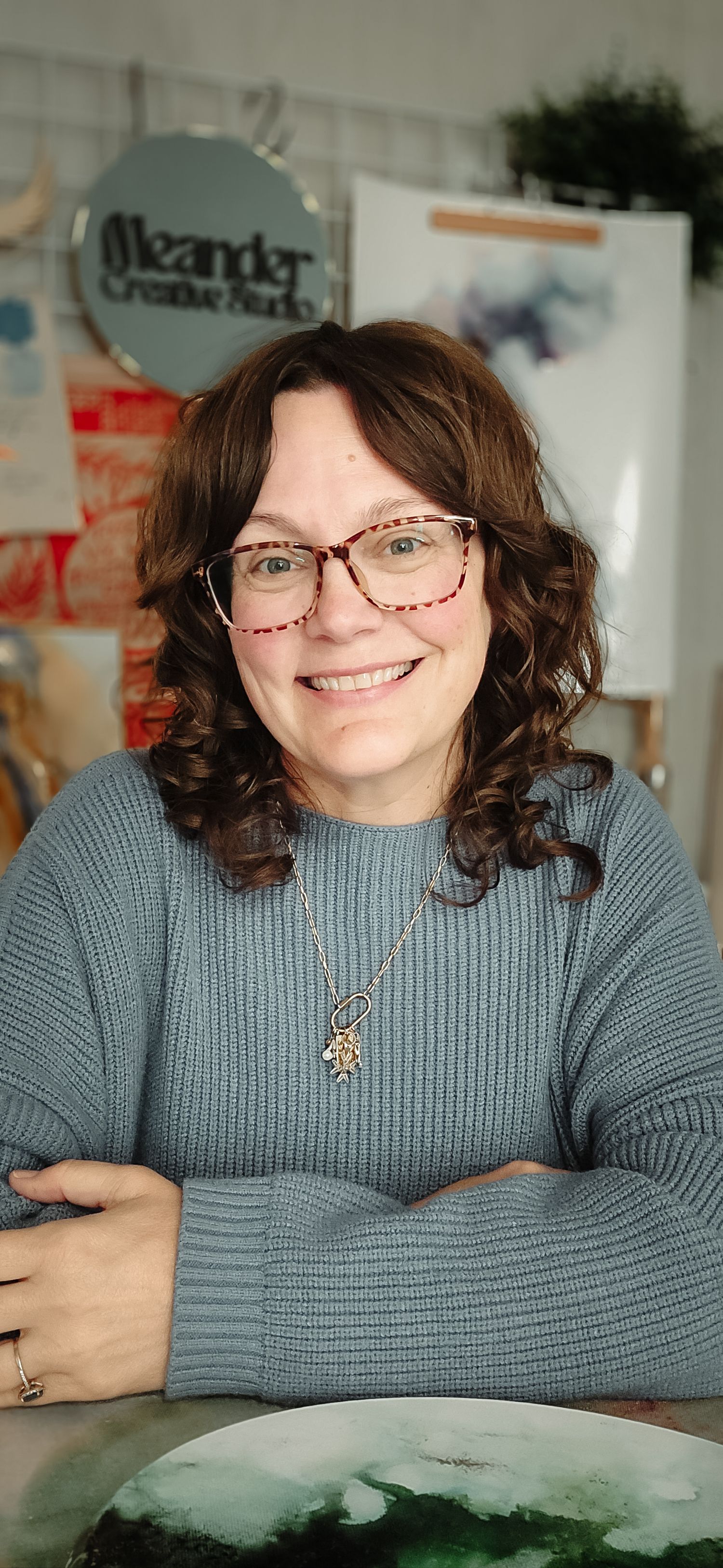 Smiling woman with curly brown hair and red glasses wearing a teal sweater and necklace, seated at Oleander Creative Studio.