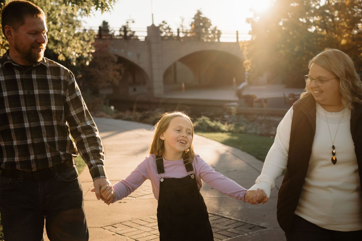 A joyful family of three holds hands and walks along a sunny park path, with a laughing girl between her smiling parents.
