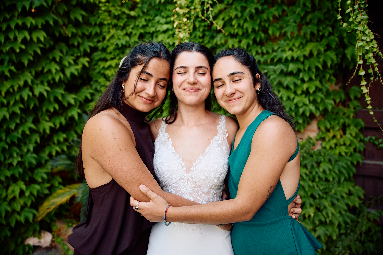 Bride embracing two wedding guests / sisters in front of a green ivy wall during her Barcelona wedding celebration.