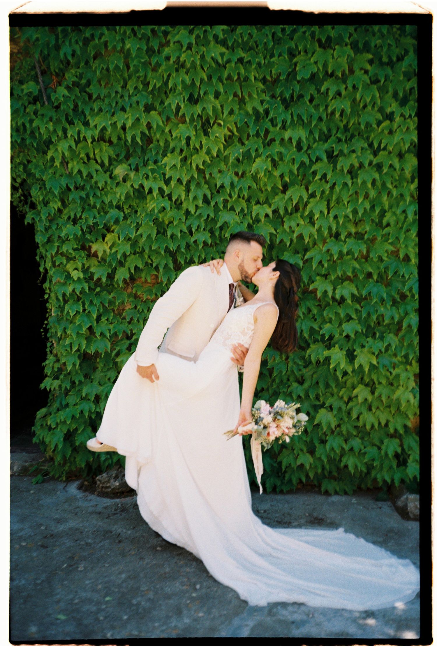 Editorial film portrait of the bride and groom kissing in front of a green ivy wall.