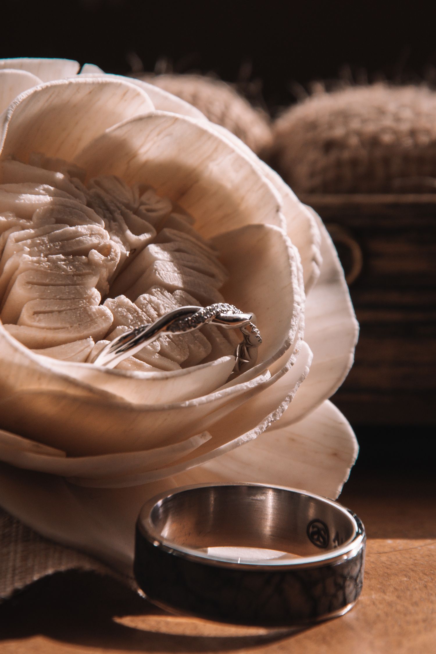 Wedding rings nestled in cream fabric rose with dark band on wooden surface in warm light.