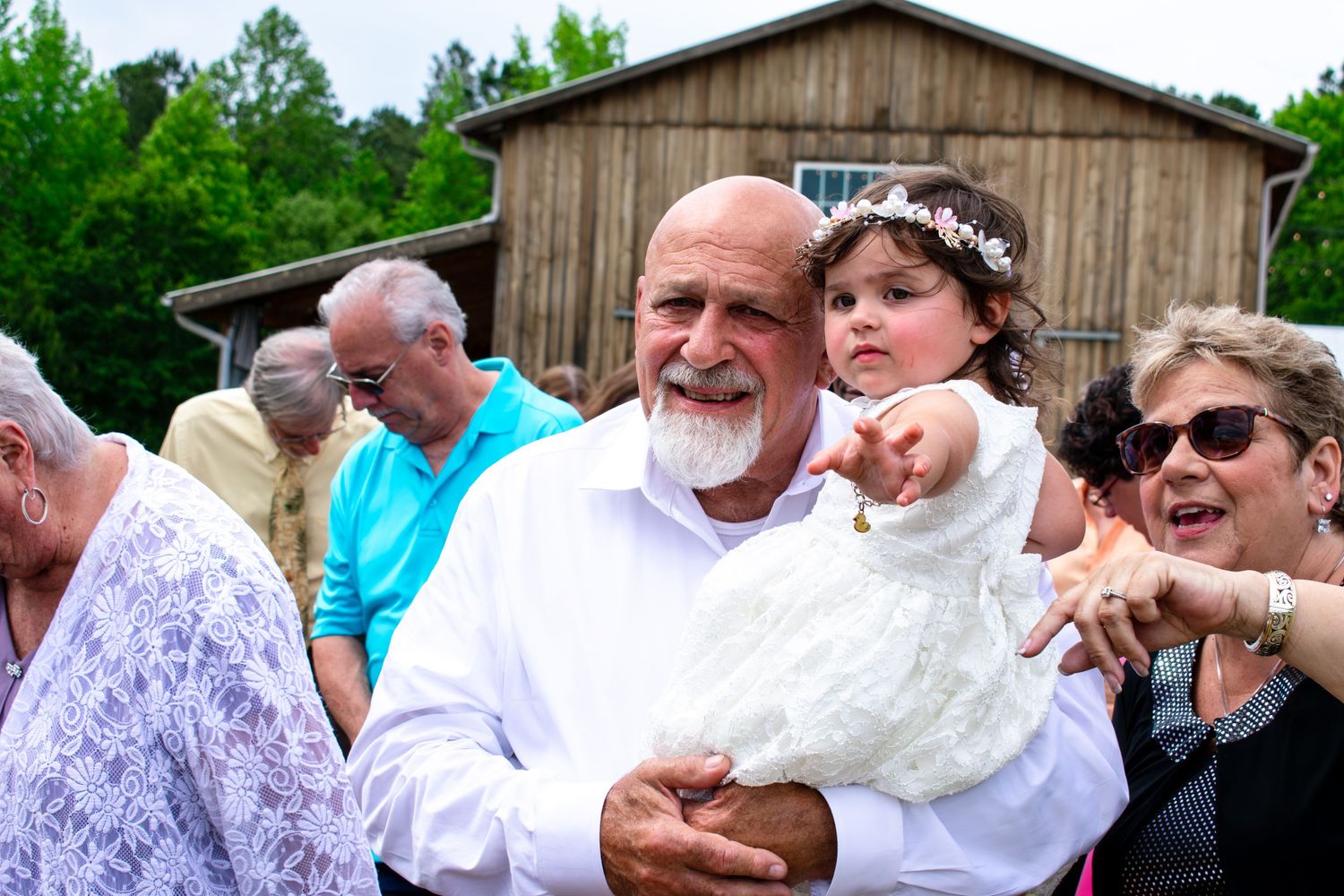 Man holding white duck with young girl in flower crown pointing at it, guests gathered outdoors near rustic barn.