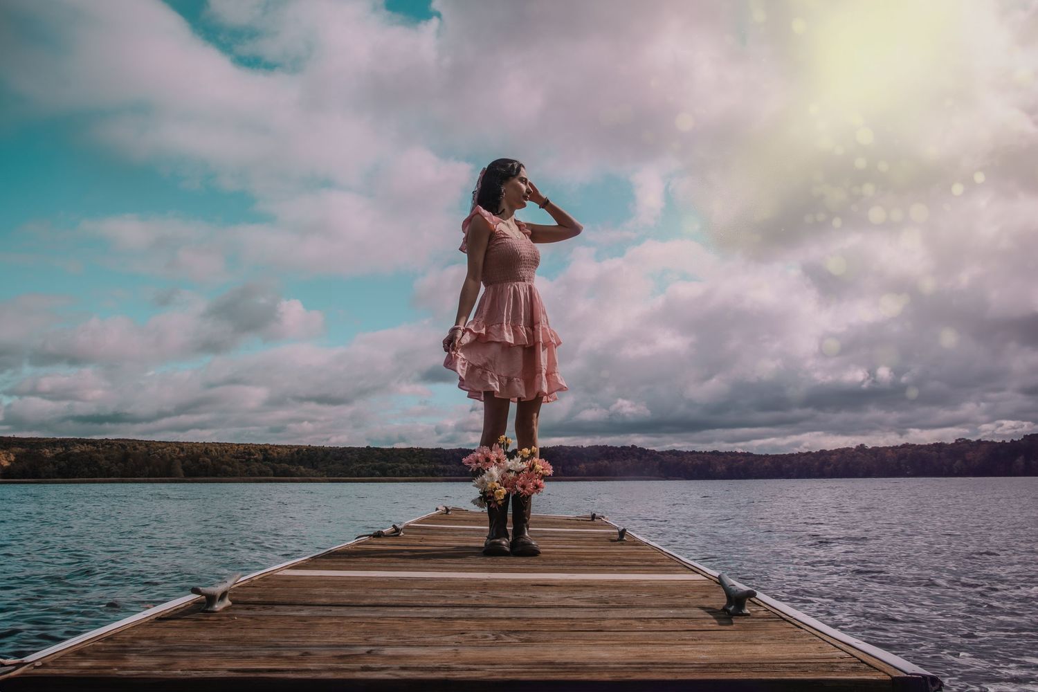 Woman in pink dress standing on wooden pier overlooking calm lake under dramatic cloudy sky.