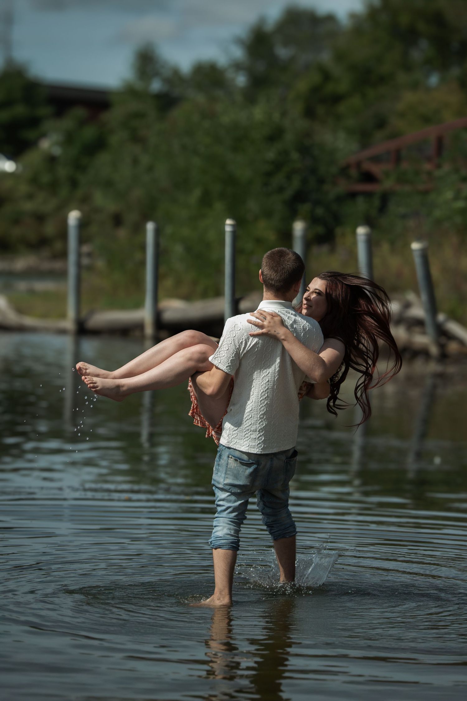 A man carries a woman in his arms while wading through shallow water near a dock on a cloudy day.