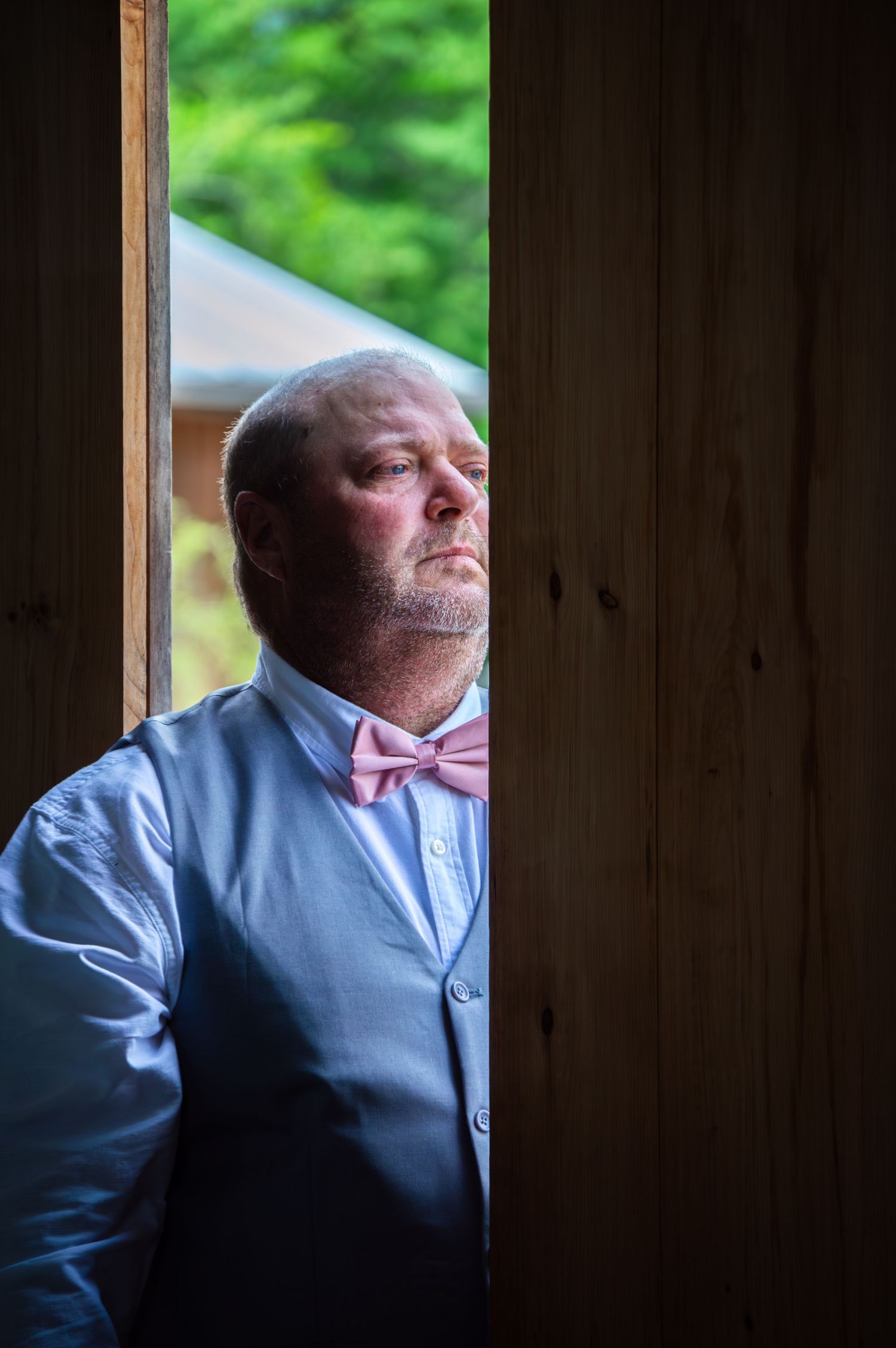 A bald man with a beard wearing a gray vest and pink bow tie gazes upward while standing in a wooden doorway.