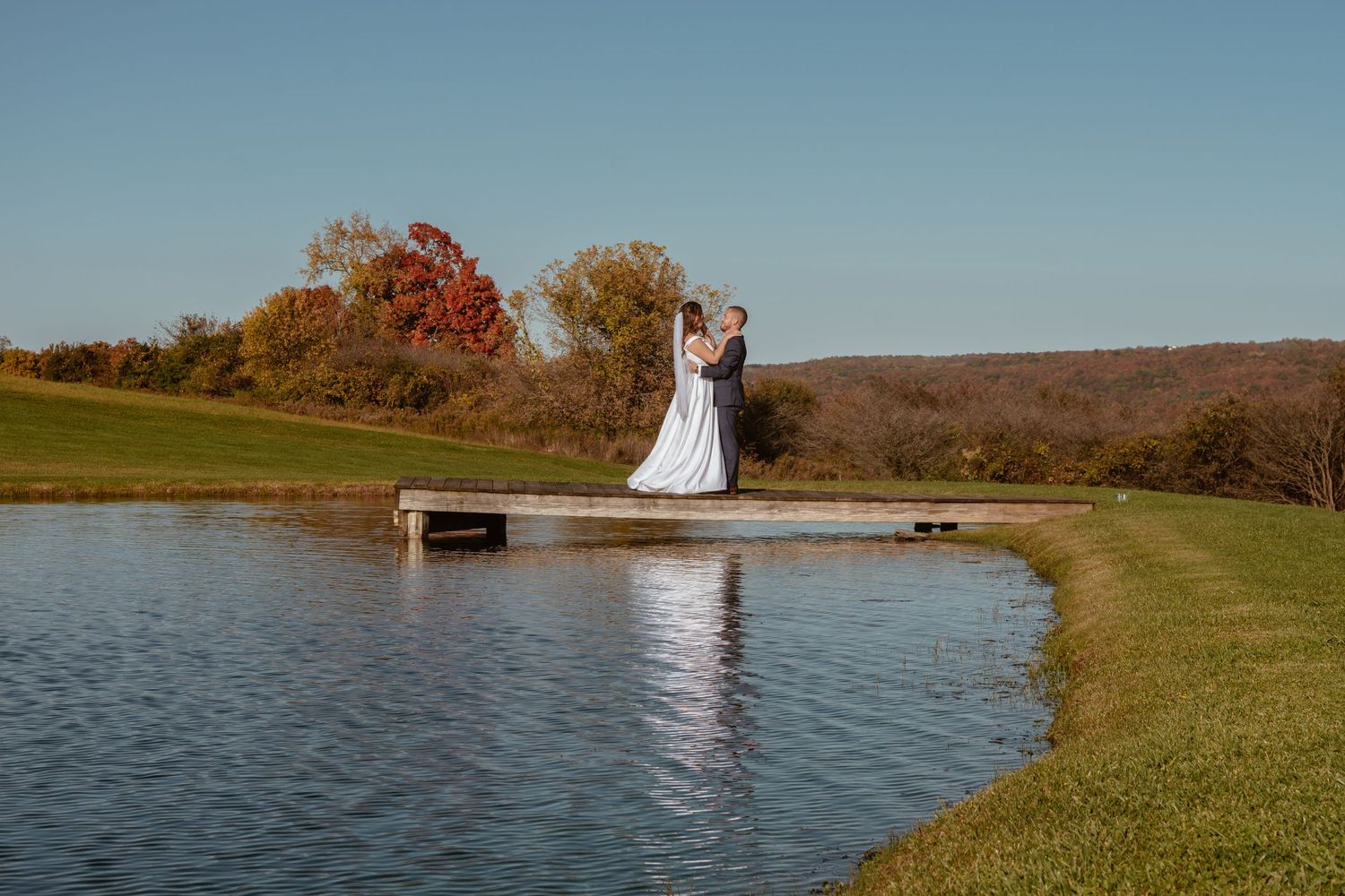 Bride and groom embrace on a dock over a calm pond, surrounded by autumn foliage and rolling hills.
