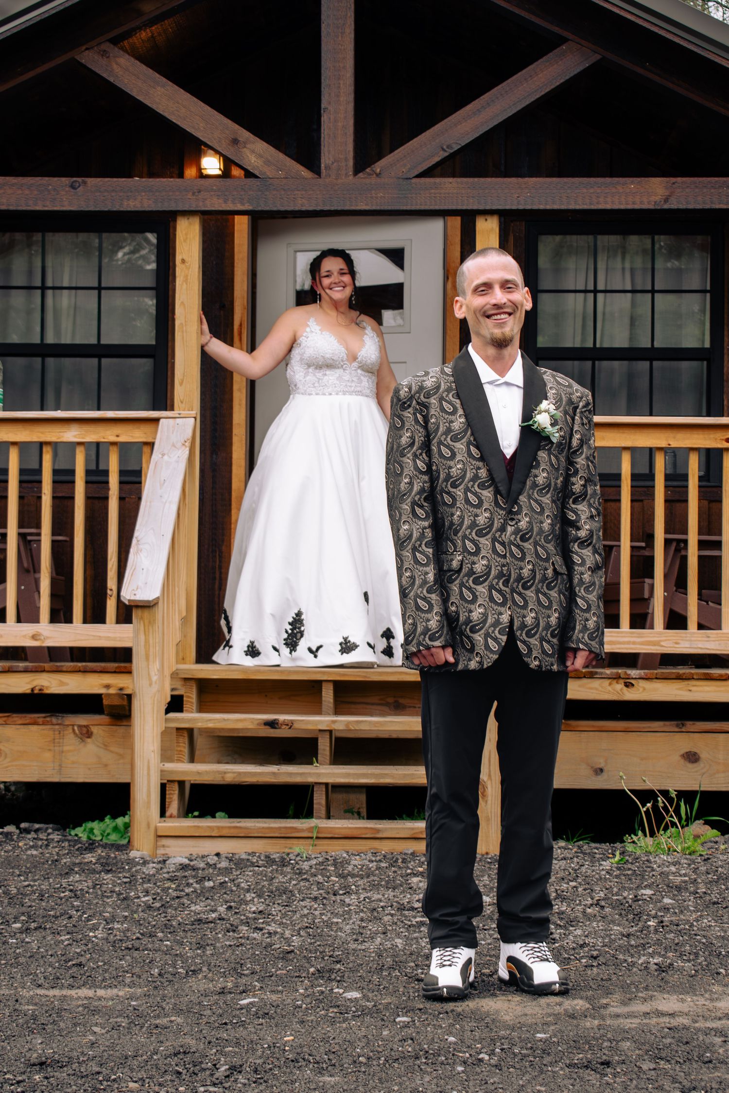 Smiling groom in ornate jacket stands before rustic cabin steps where bride in white gown poses on the porch.
